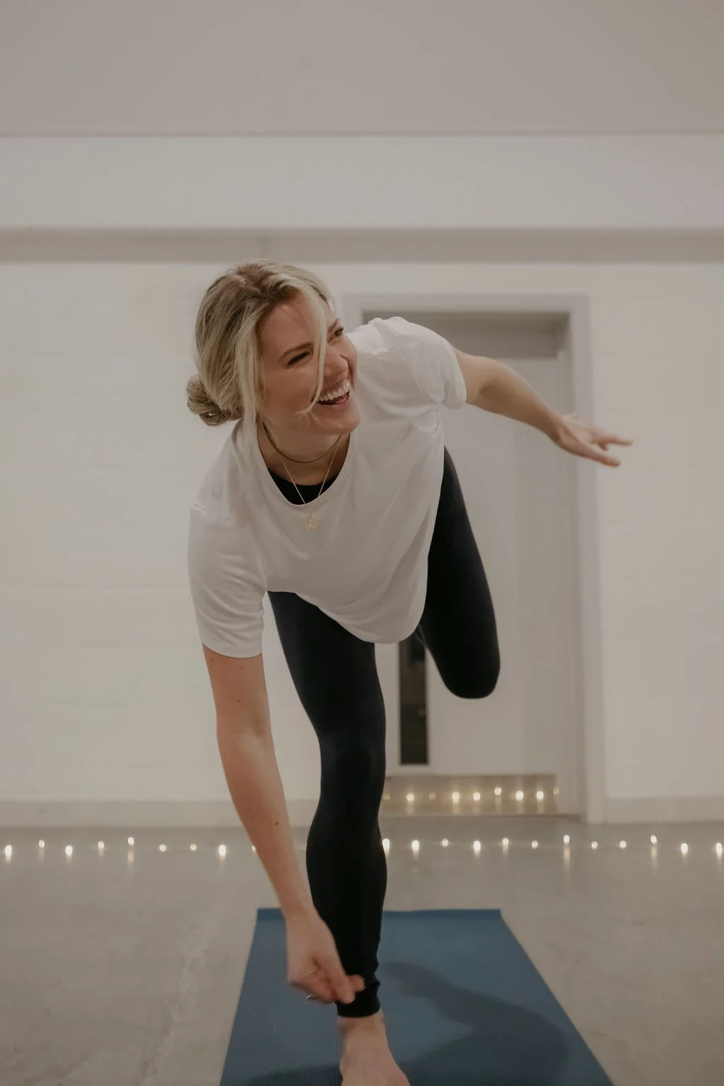 Woman practicing yoga indoors on a blue mat, smiling while balancing on one leg and holding her other foot behind her.