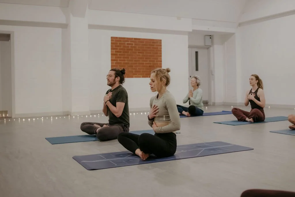 Four people practicing yoga on mats in a calm, minimal studio with white walls, a brick accent wall, and string lights along the floor.