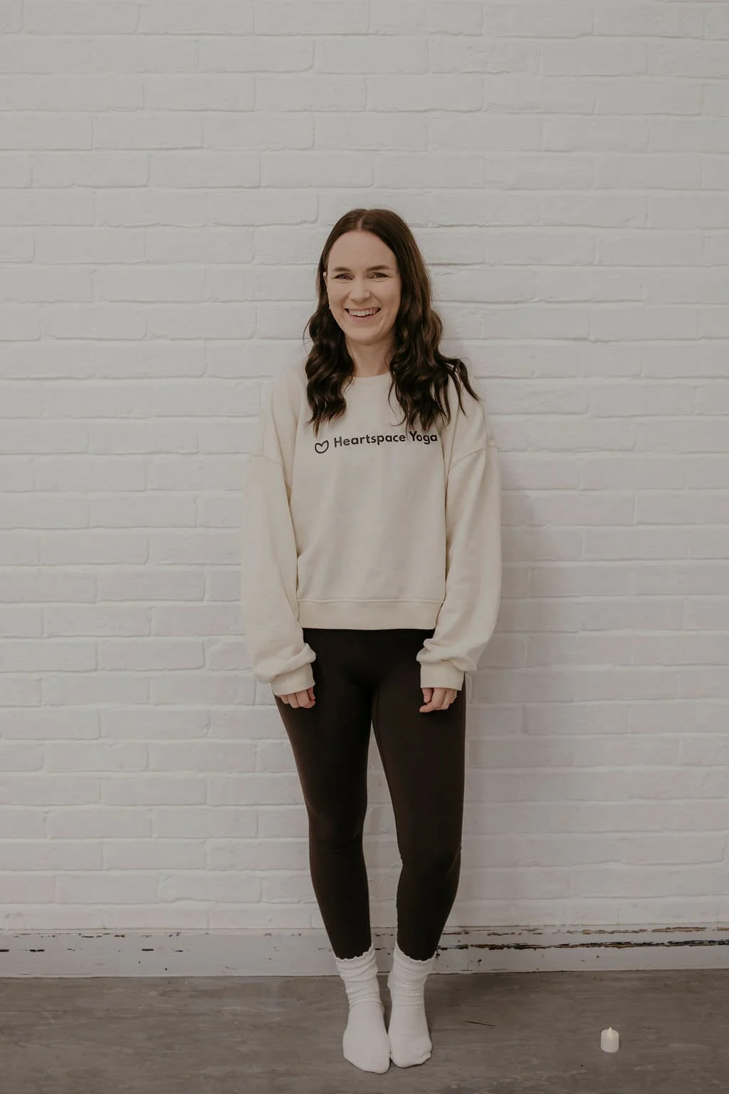 Woman with brown hair smiling, wearing a cream sweatshirt with 'Heartspace Yoga' logo, black pants, and white socks, standing against a white brick wall with a small candle on the floor.