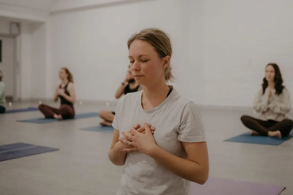 A woman practicing meditation with her eyes closed and hands on her chest, seated on a yoga mat in a room with other women meditating in the background.
