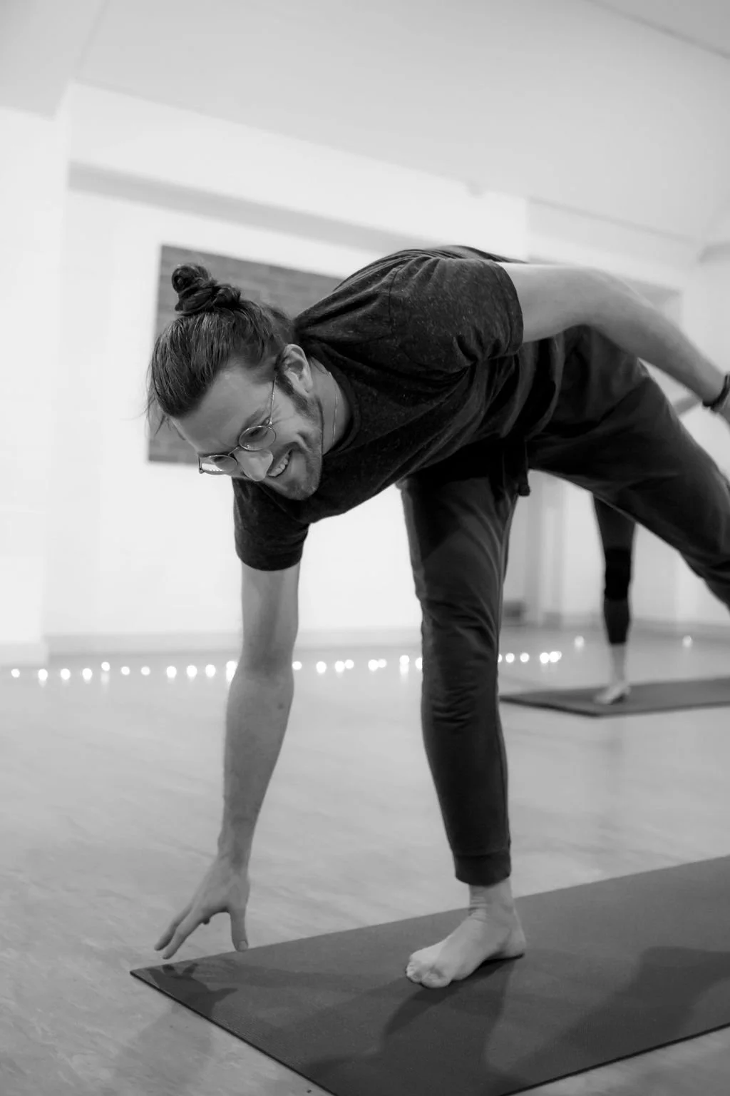 A man with glasses and a man bun practicing yoga on a mat, smiling, in a bright room with string lights and a wall decoration in the background.