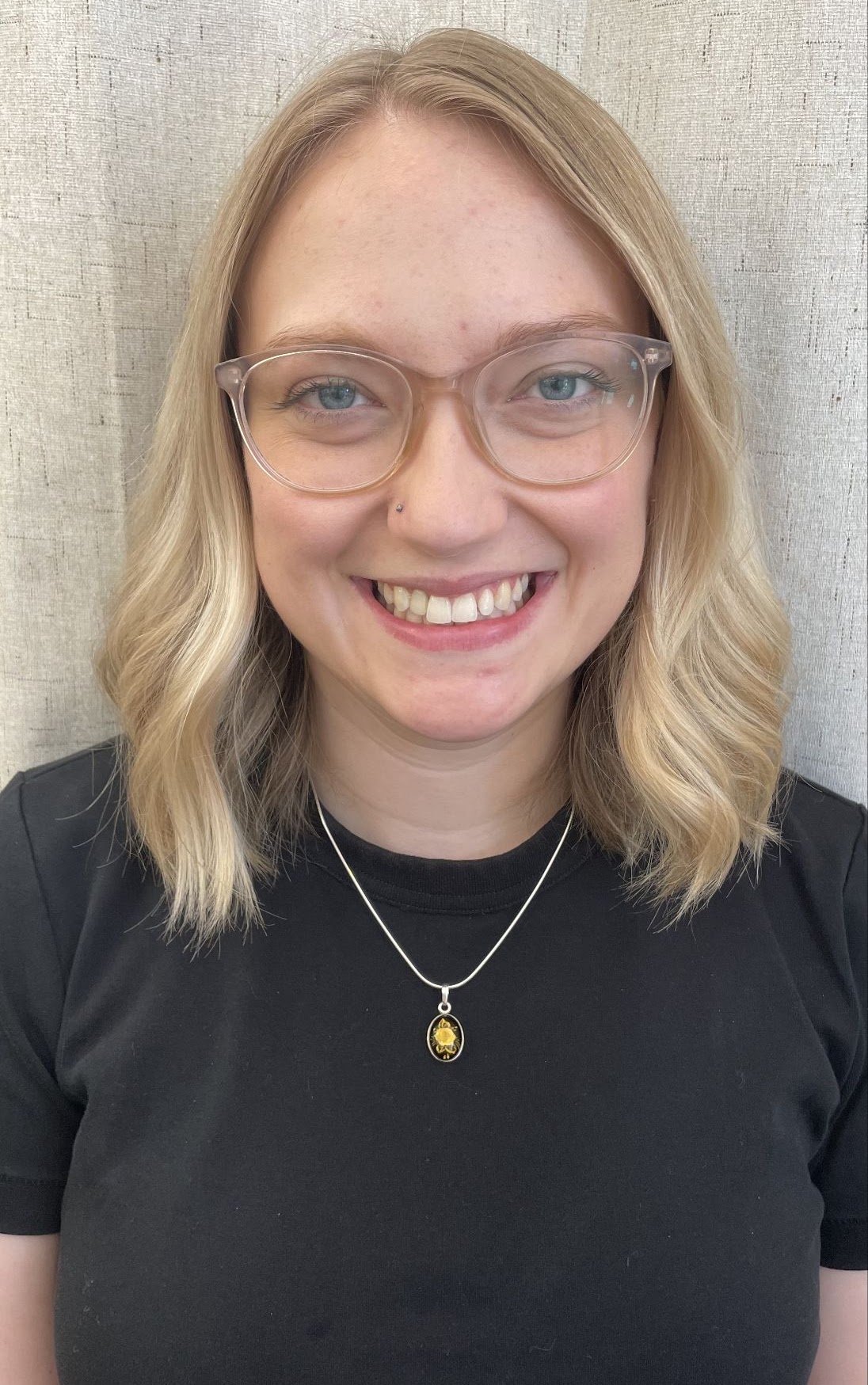 A young woman with blonde shoulder-length hair, wearing glasses, a black T-shirt, a silver necklace with a gold pendant, and smiling showing her teeth, standing in front of a light-colored textured background.