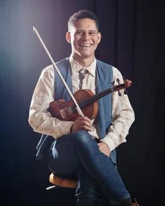 Young man smiling, holding a violin and a bow, sitting in front of a dark background.