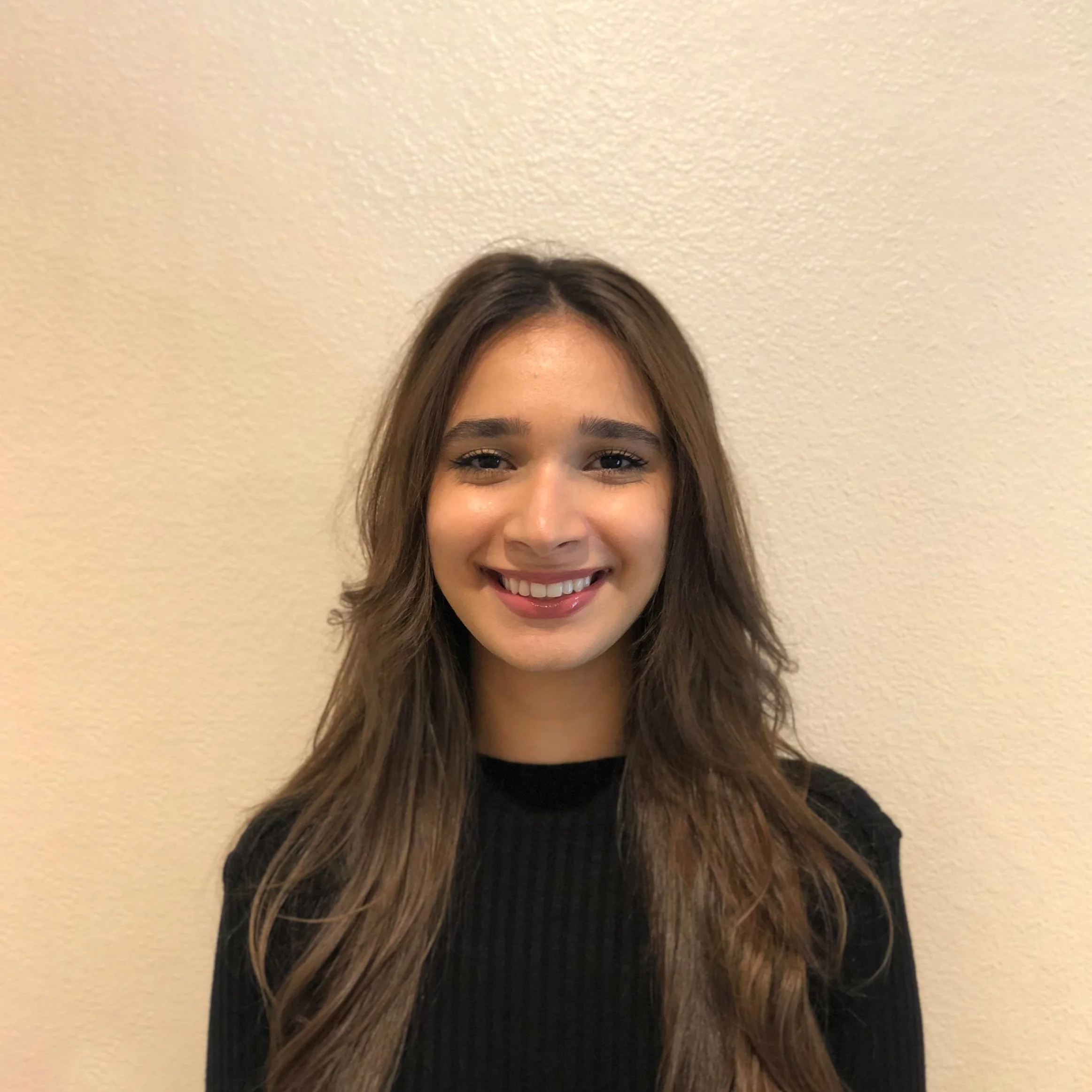 Young woman with long wavy brown hair smiling, wearing a black top, standing against a plain beige wall.