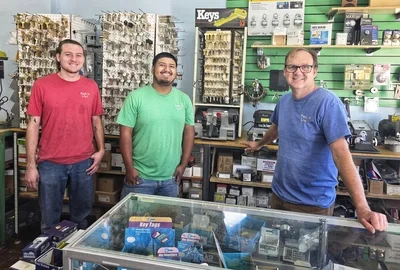 Three locksmiths behind the counter of the Payson Lock & Key store in Utah, with keys and locks displayed on the wall behind them.