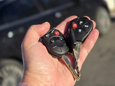 Hand holding a backup car key with two key fobs, one with buttons for locking and unlocking doors, on a blurred outdoor background in Payson, Utah.
