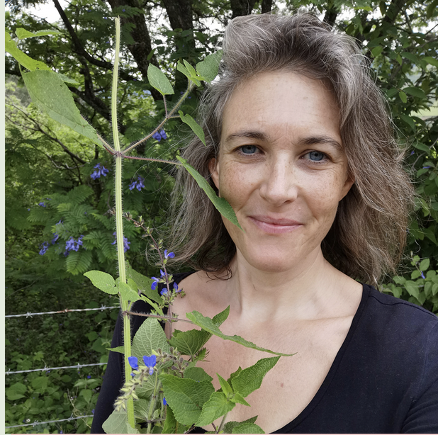 A woman in a lush green garden with a tall, leafy plant with blue flowers in the foreground.