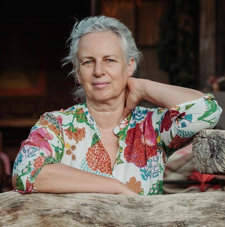 An older woman with gray hair, wearing a colorful floral blouse, resting her hand on the back of her neck, sitting outdoors with a rustic wooden background.