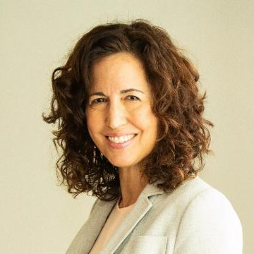 A woman with curly brown hair smiling, wearing a light-colored blazer, against a neutral background.