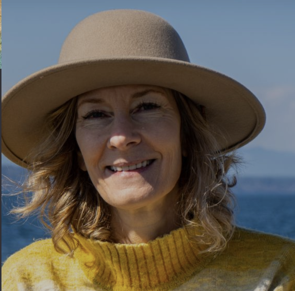 A woman with wavy hair wearing a large beige hat and a yellow sweater, smiling outdoors with water and sky in the background.