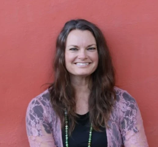Woman with long brown hair smiling, wearing a purple patterned jacket and a necklace, standing against a red wall.
