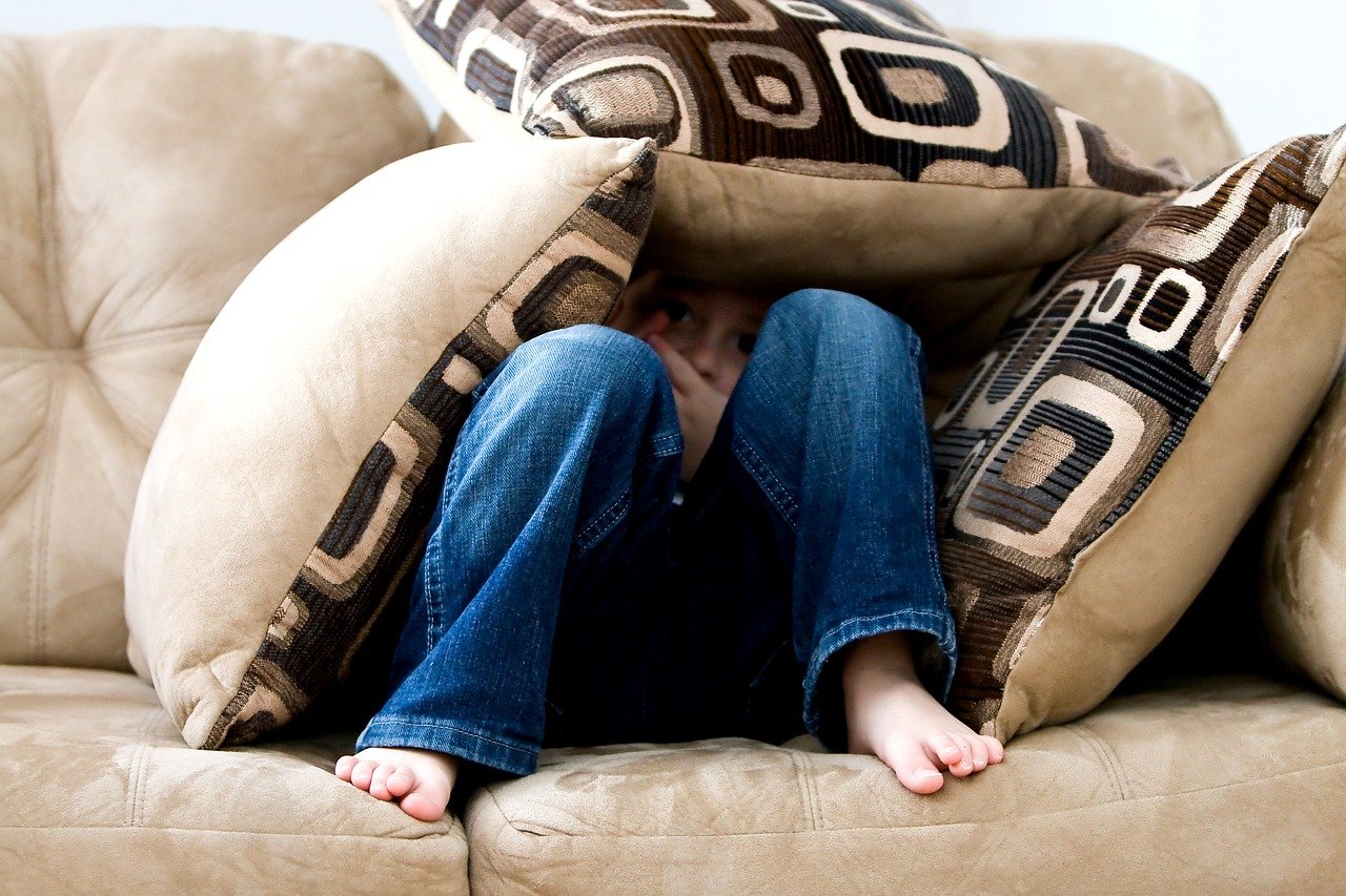 A young child hiding behind and surrounded by large decorative pillows on a beige couch.