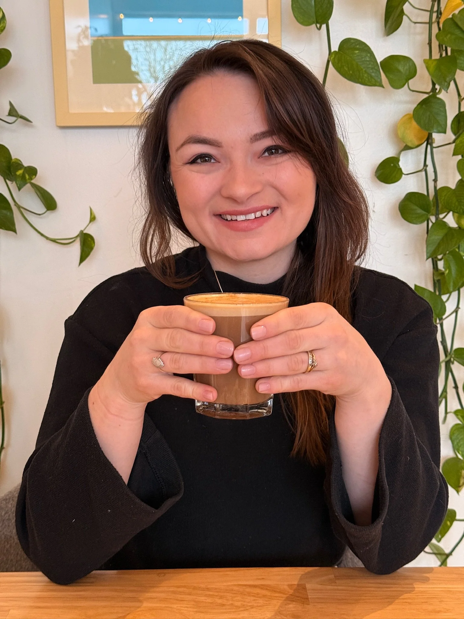 A woman with shoulder-length brown hair holding a glass of hot cocoa with whipped cream, smiling at the camera, seated at a wooden table with green plants and a framed picture in the background.