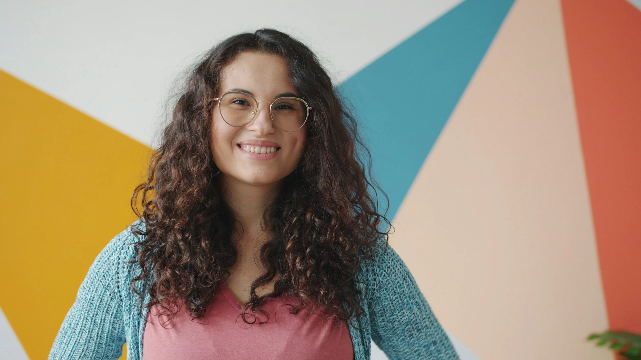 A woman with curly brown hair and glasses smiling, standing in front of a colorful geometric mural.