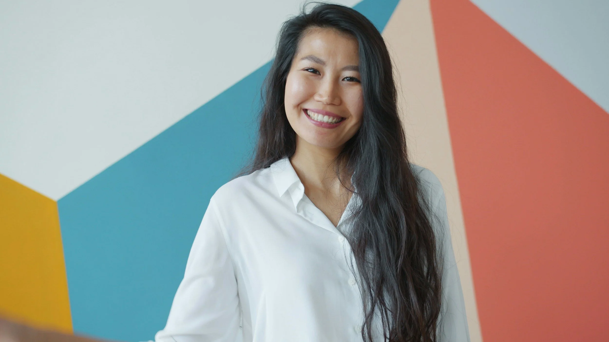 A smiling woman with long black hair wearing a white shirt standing in front of a colorful geometric mural.