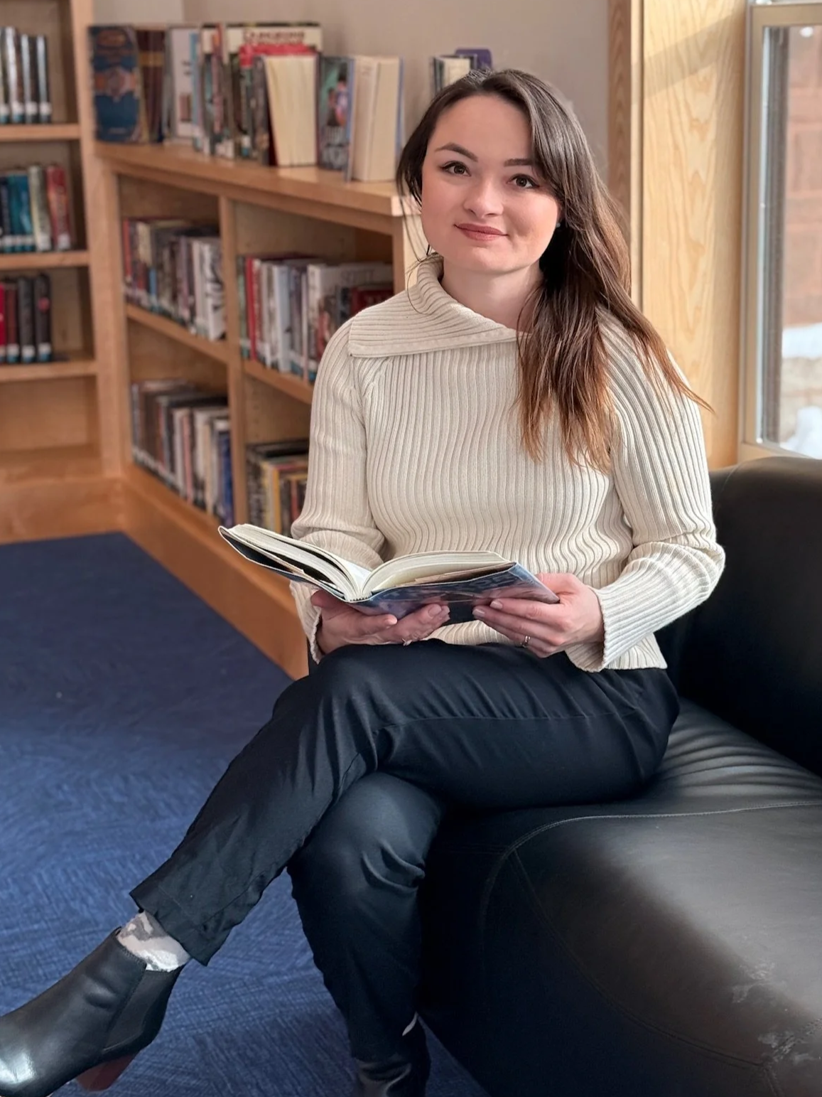 A young woman with long brown hair sitting on a black leather couch in a library, holding an open book, wearing a cream ribbed sweater and black pants.
