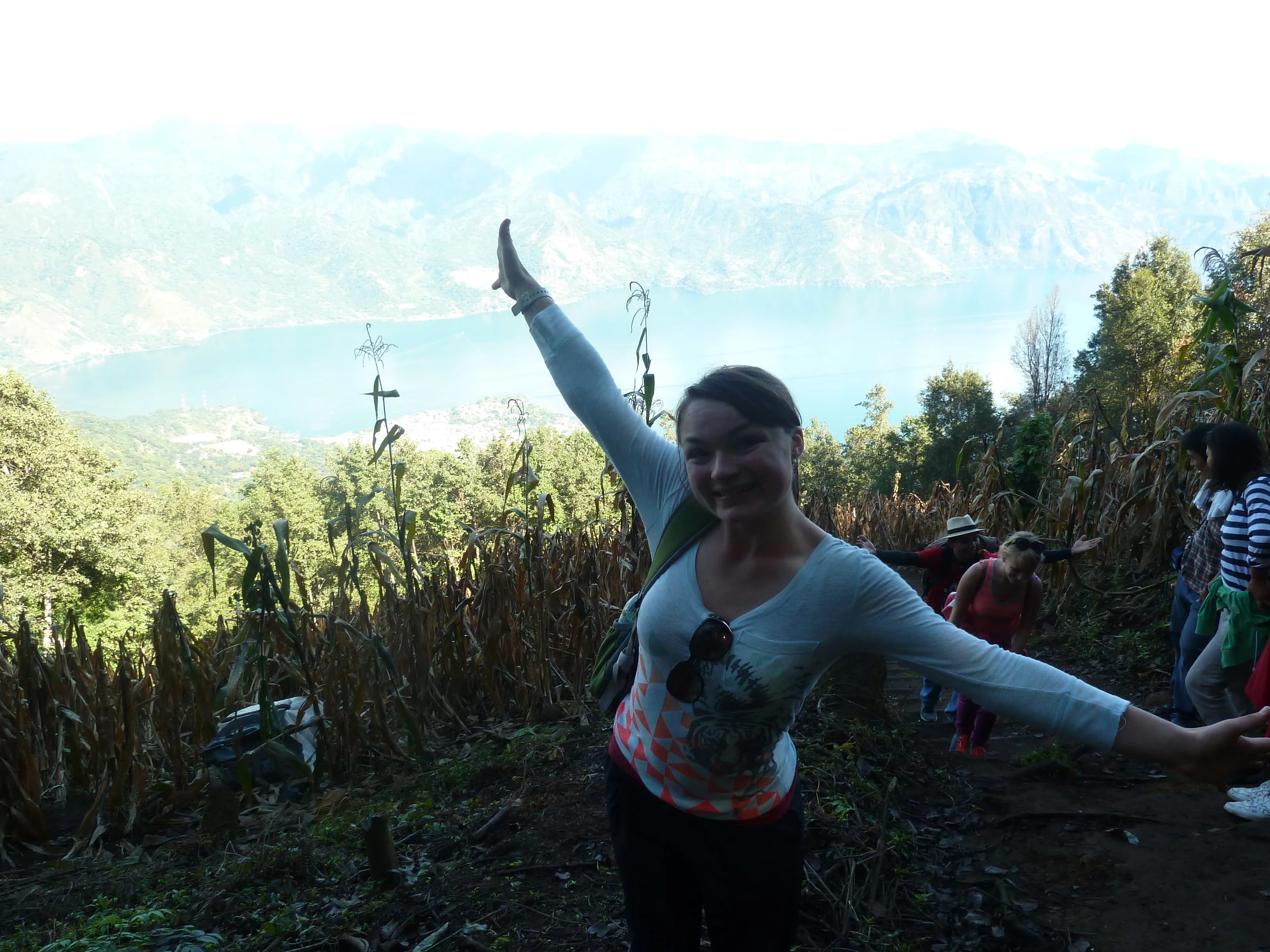A group of people hiking on a trail through a cornfield with a lake and mountains in the background. The woman in the foreground is smiling and gesturing with her arms outstretched.