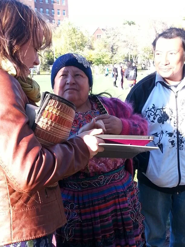 Three people standing outdoors in a park, engaged in conversation. One woman is holding a tablet, and the other woman is wearing traditional clothing and a blue knit hat. A man stands nearby, looking at the women.