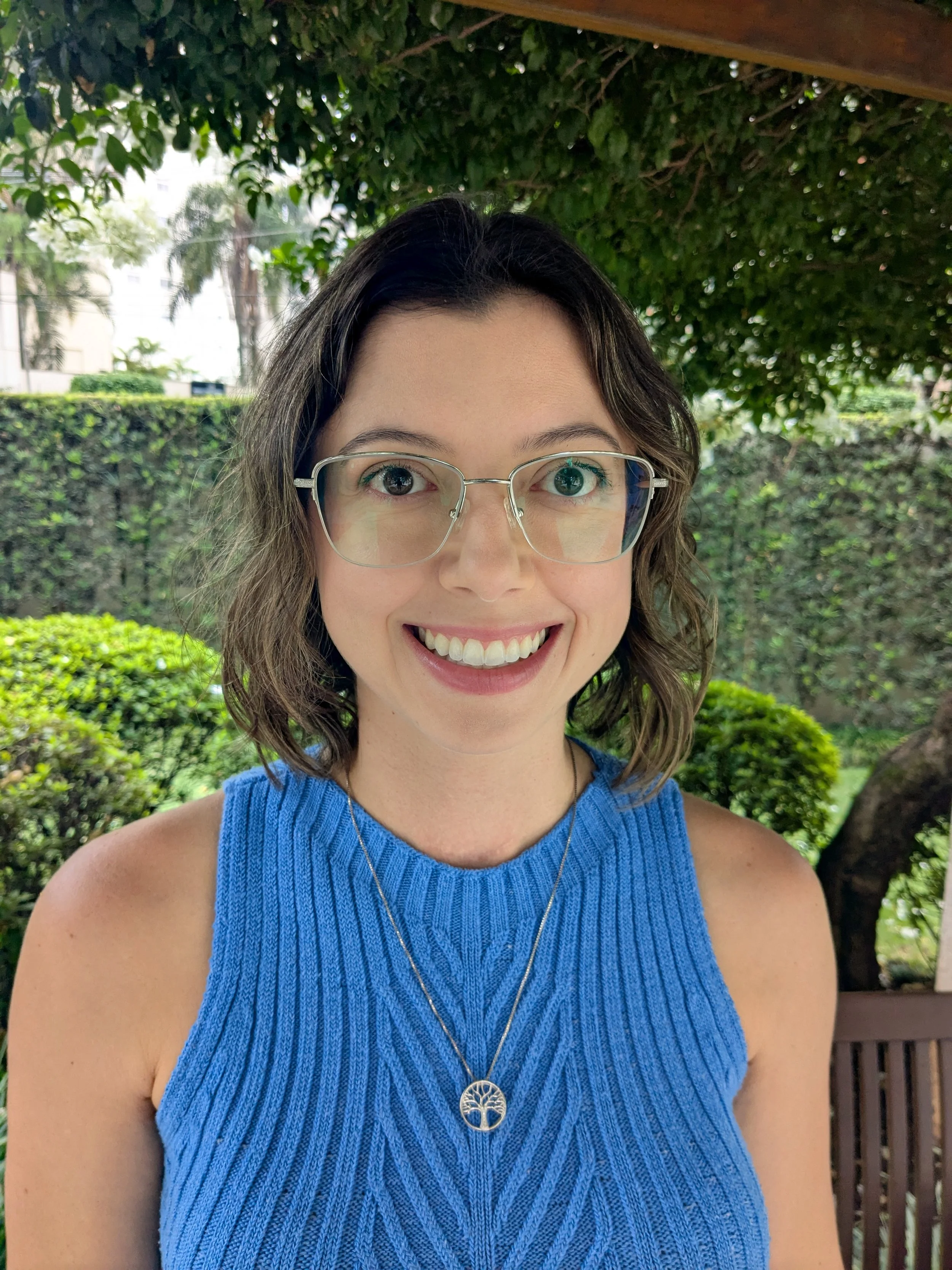 A young woman with shoulder-length wavy brown hair, wearing clear glasses, a blue sleeveless knit top, and a silver necklace with a tree pendant, smiling outdoors with green foliage and trees in the background.
