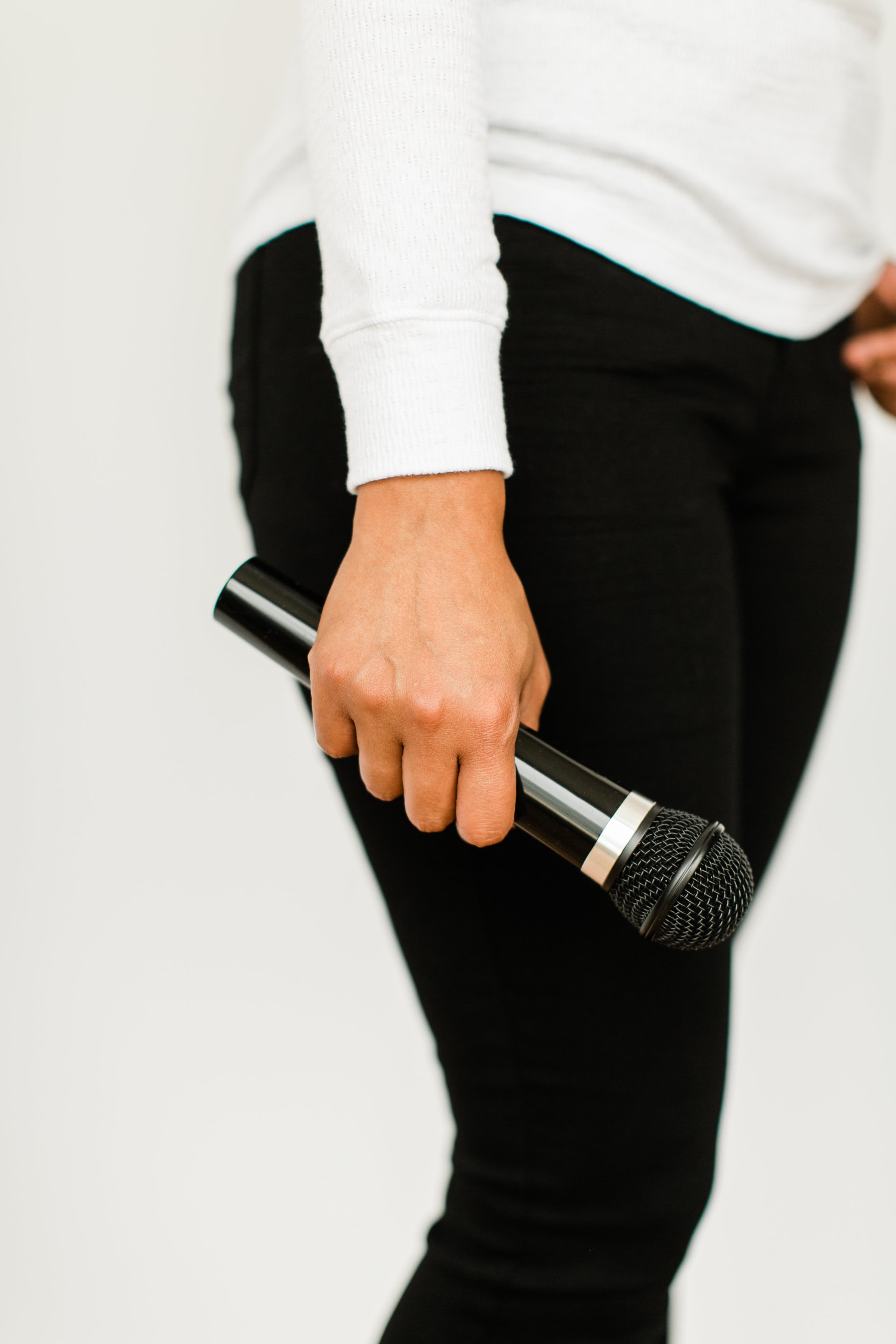Person holding a microphone by their side, wearing a white long-sleeve top and black pants.