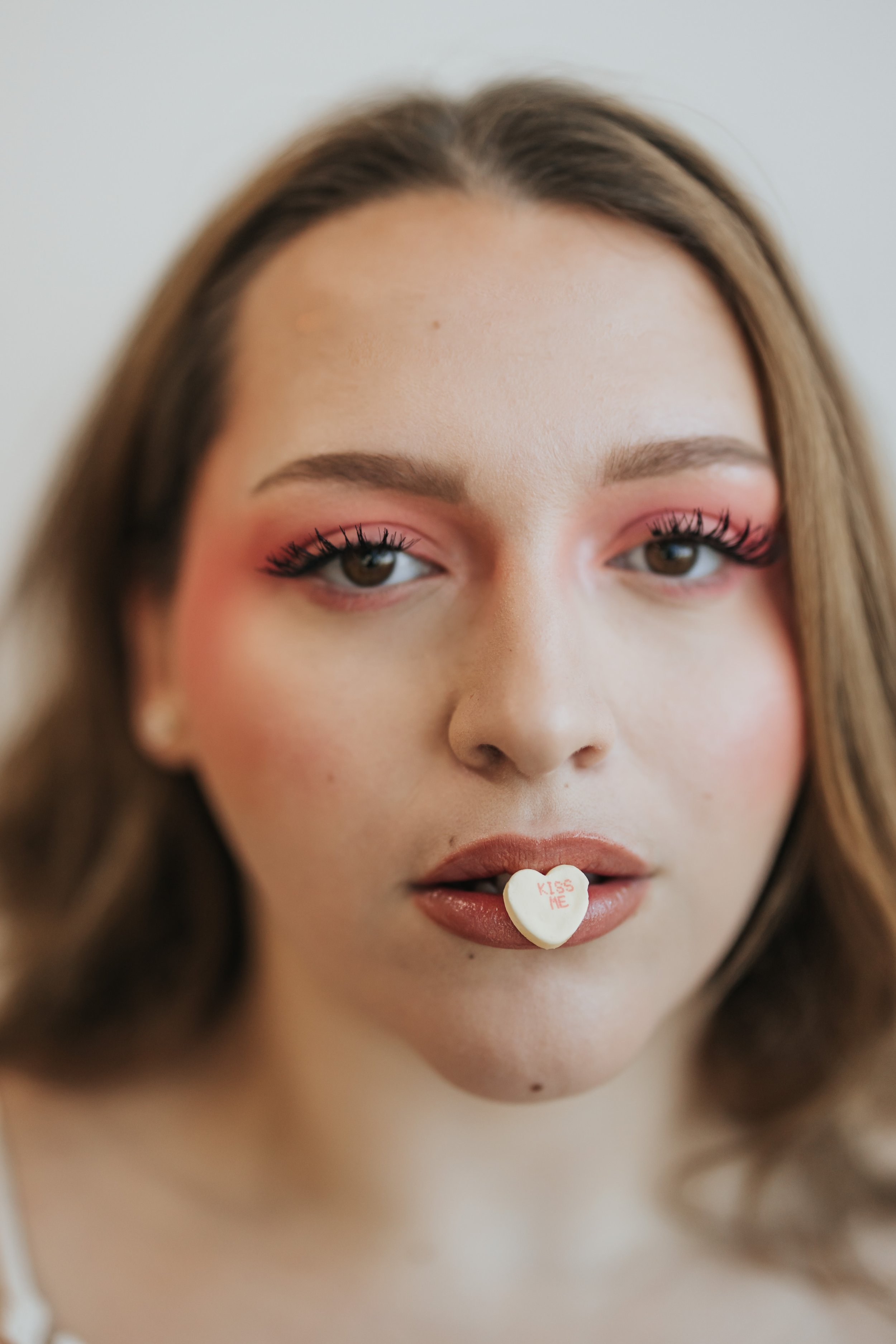  Close up photo of model with pink makeup and a candy heart in her mouth for a creative Valentine’s Day fashion photoshoot. 