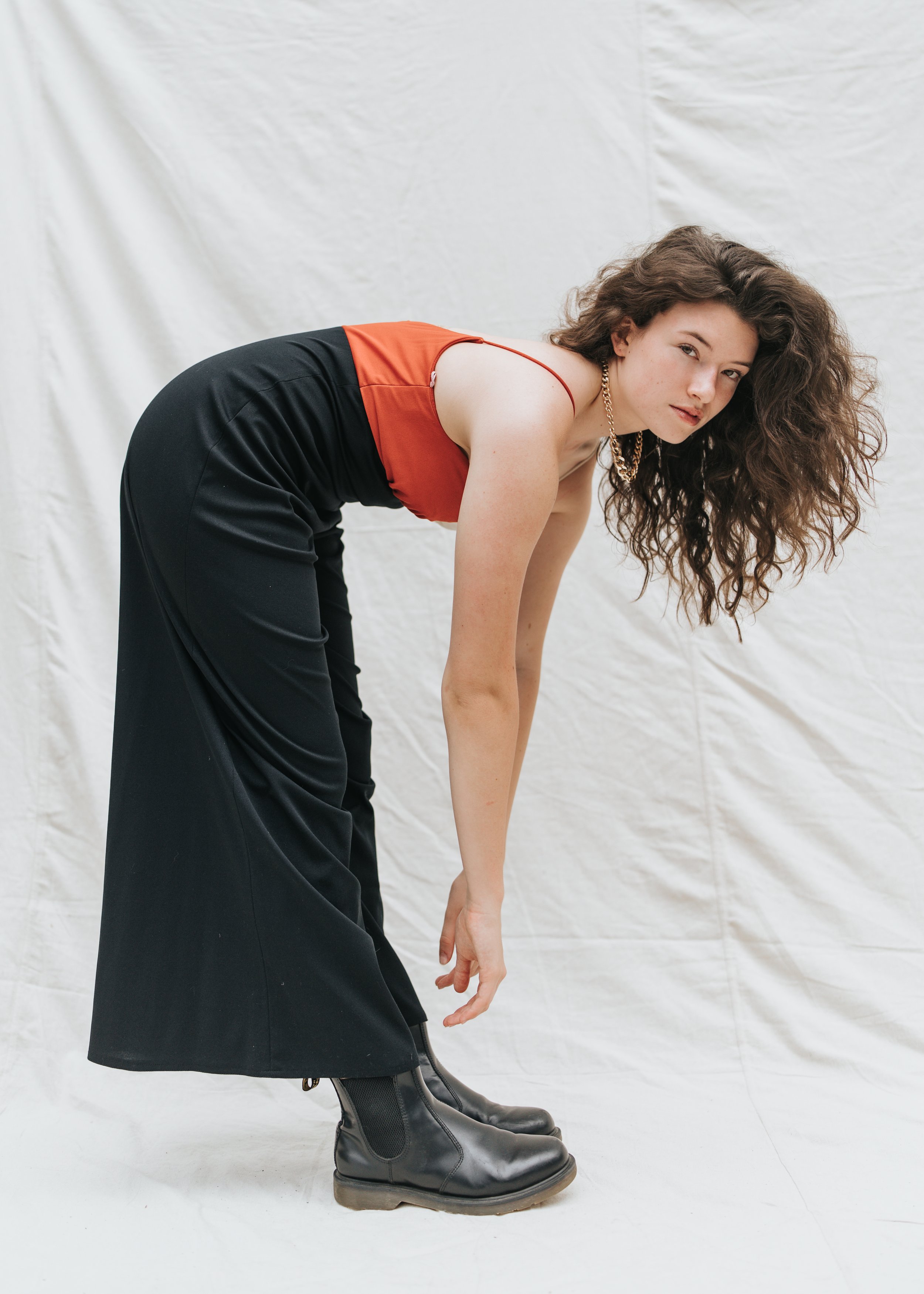  Brunette model leans over and poses for a fashion test shoot wearing black pants and an orange shirt. 