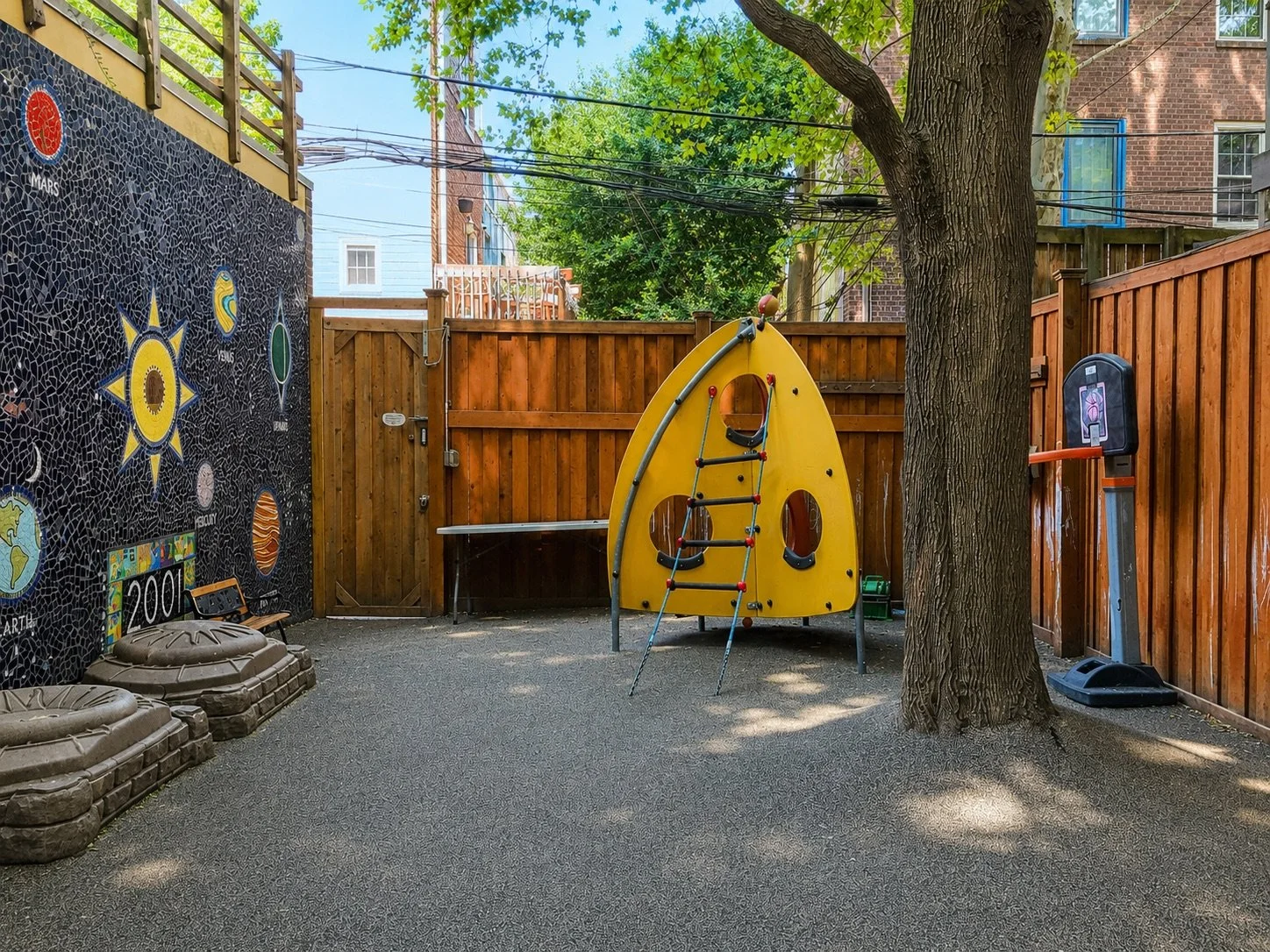 Shaded preschool playground with climbing equipment, a basketball hoop, mature trees, and a solar-system mosaic