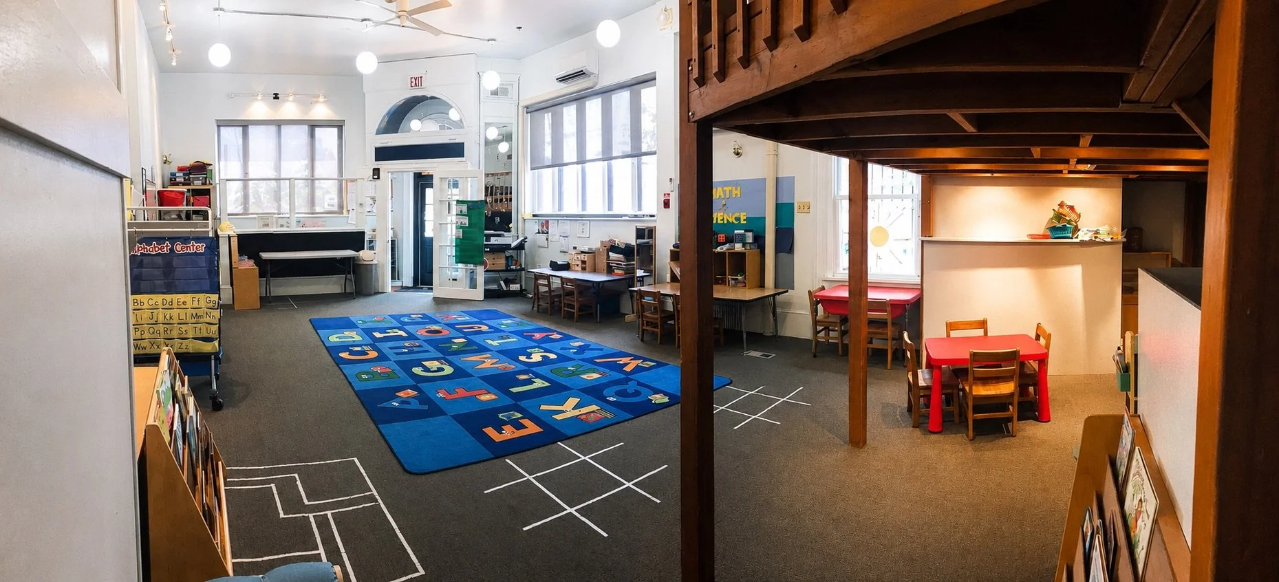 Spacious play-based preschool classroom with an alphabet rug, activity tables, and a wooden learning loft