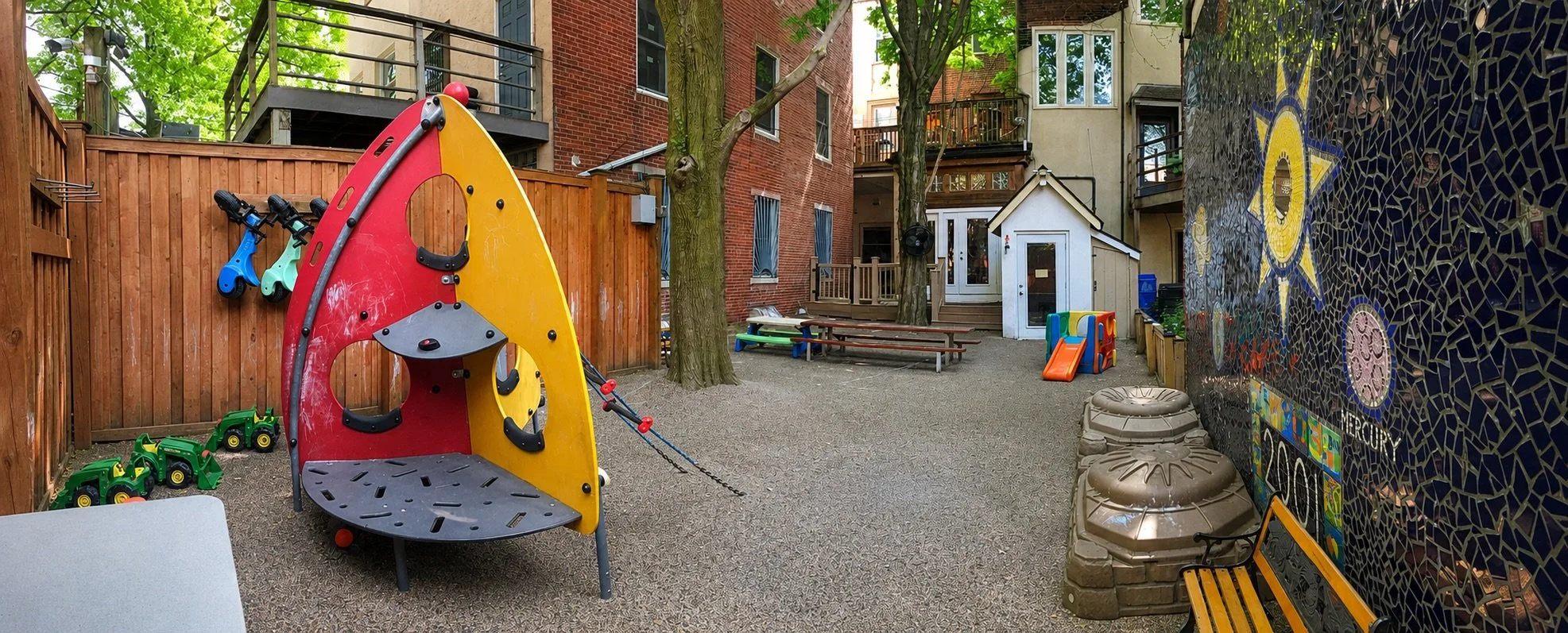 Enclosed outdoor preschool play area with climbing equipment, ride-on toys, picnic tables, and a colorful mosaic wall