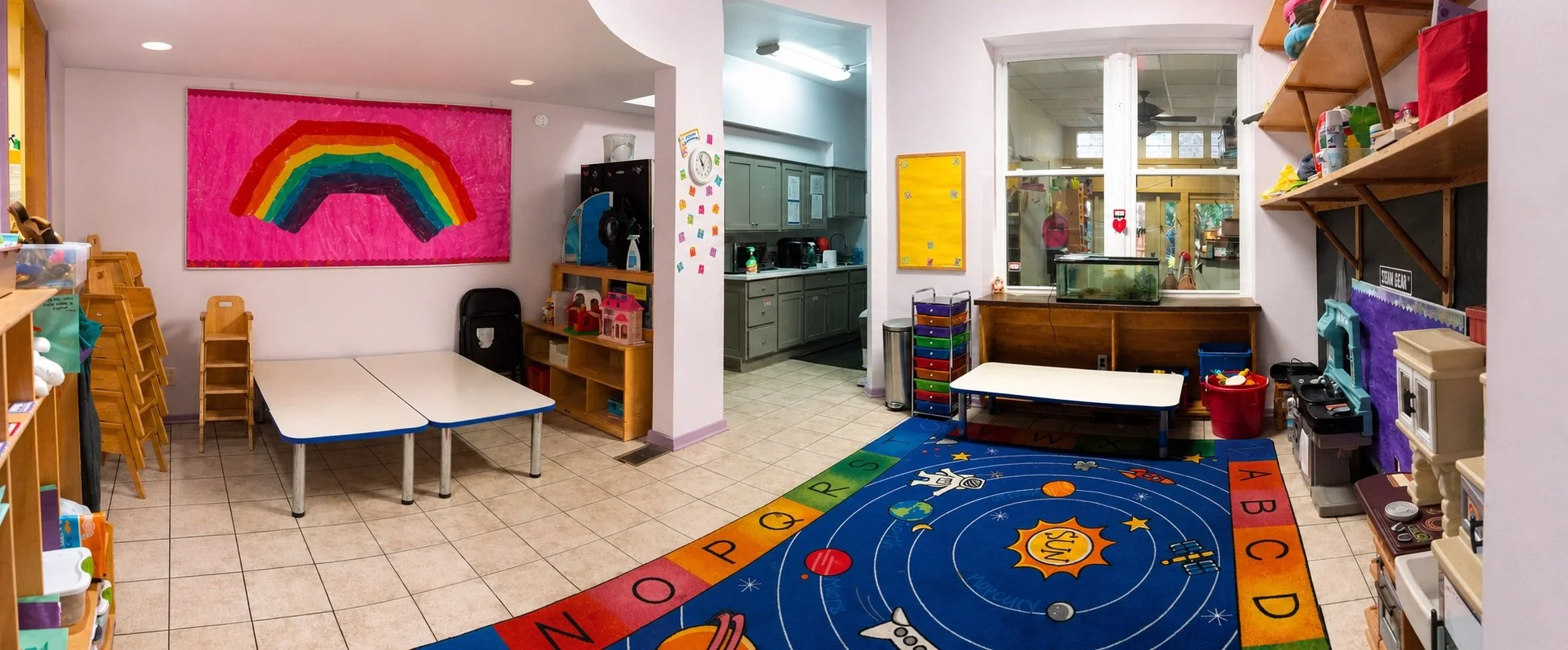 Play-based learning classroom with activity tables, dramatic play toys, and a colorful educational rug