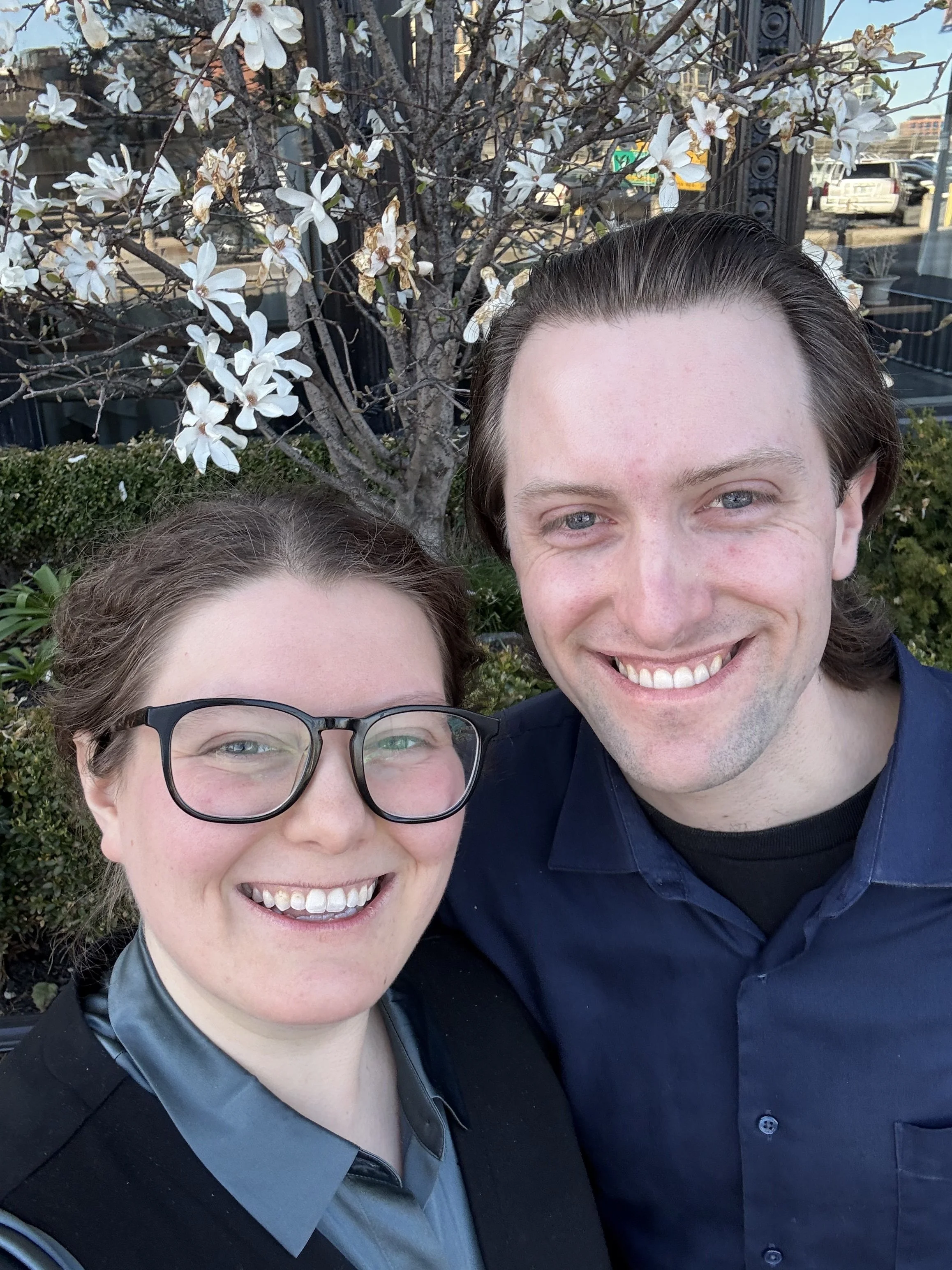 Close-up of two smiling people, a woman with glasses and a man with medium-length hair, outdoors near a flowering tree and some parked cars.