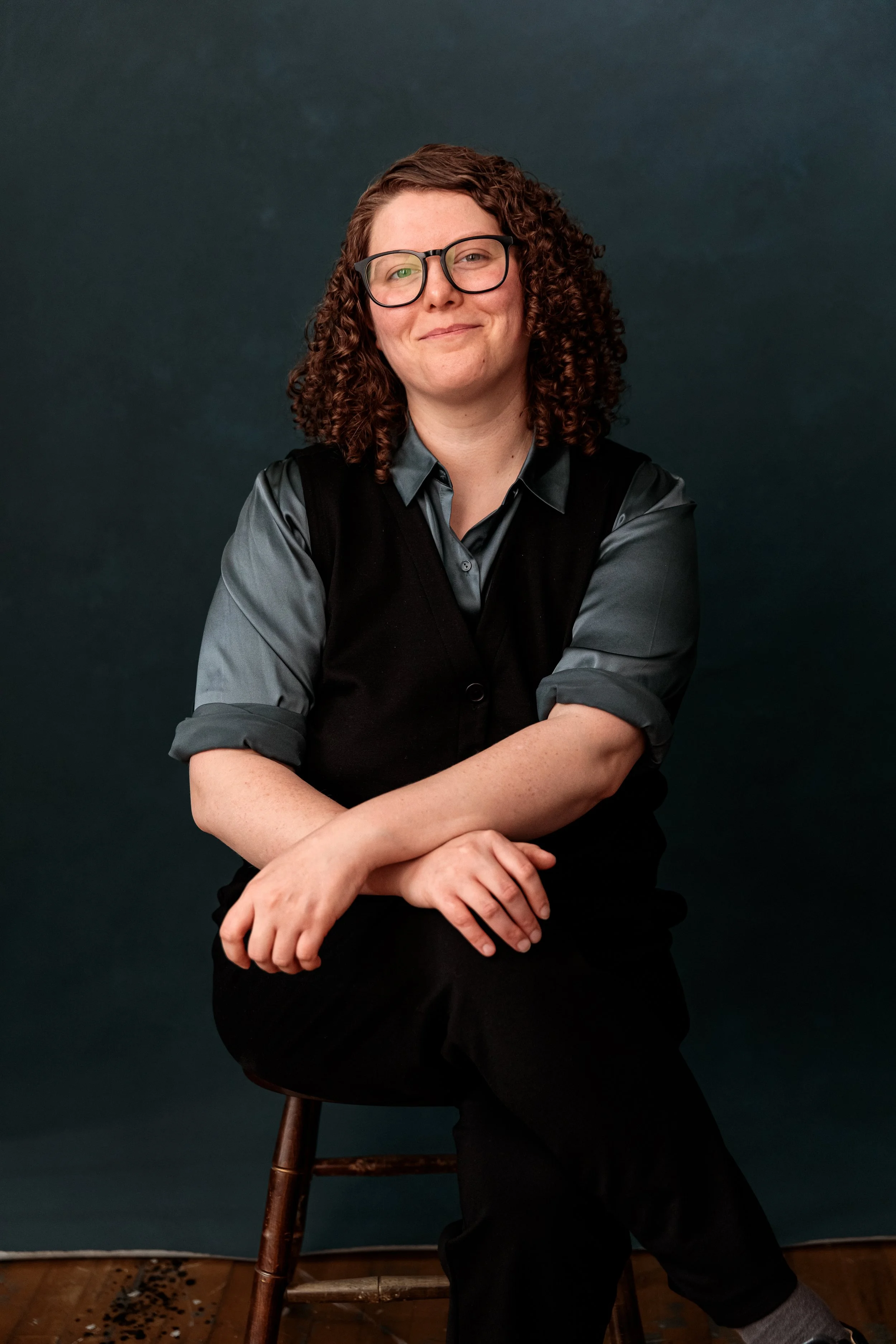 A woman with curly brown hair and glasses sitting on a wooden stool against a dark background, wearing a gray shirt and black vest.