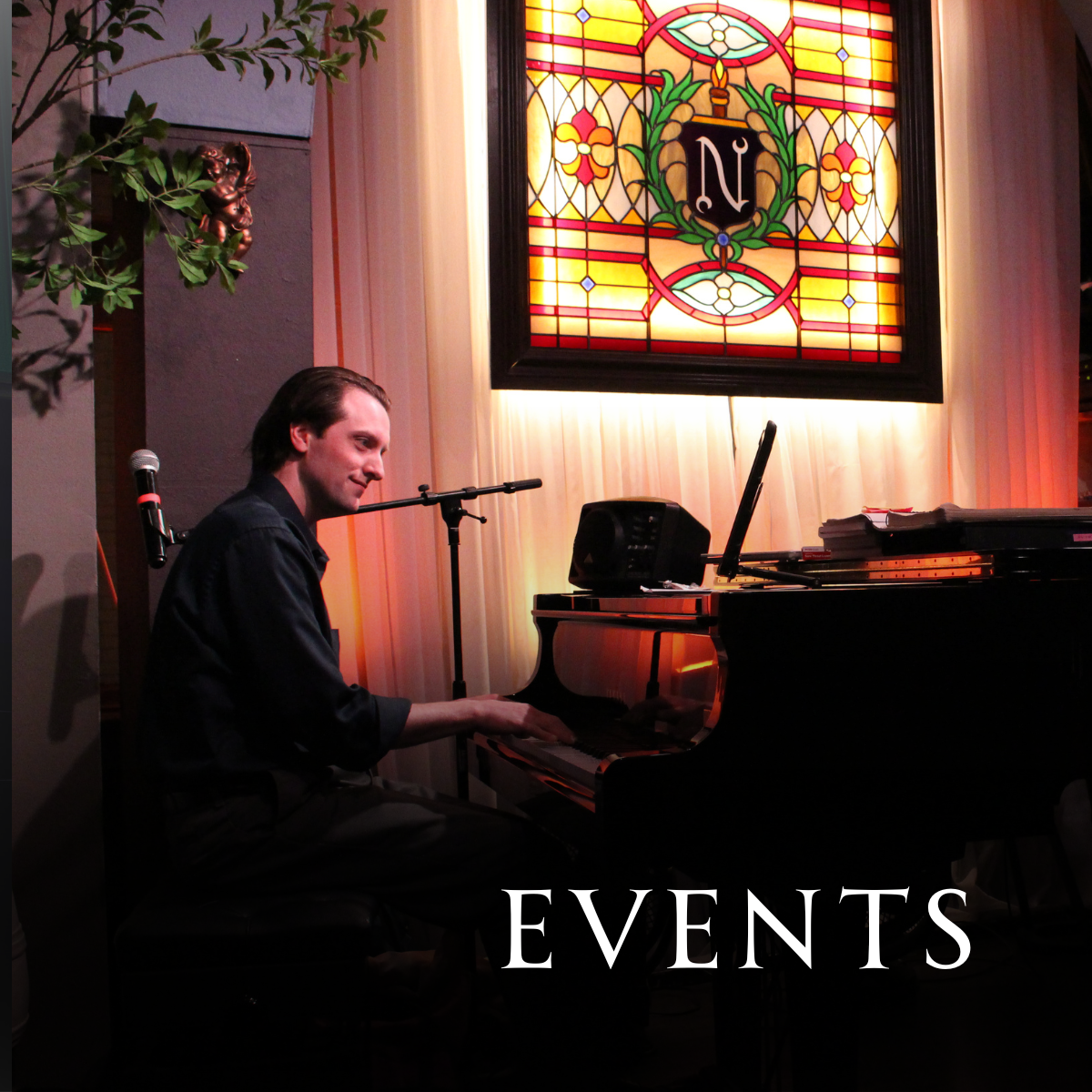 A man playing a grand piano during a live music event in a venue with stained glass artwork on the wall.