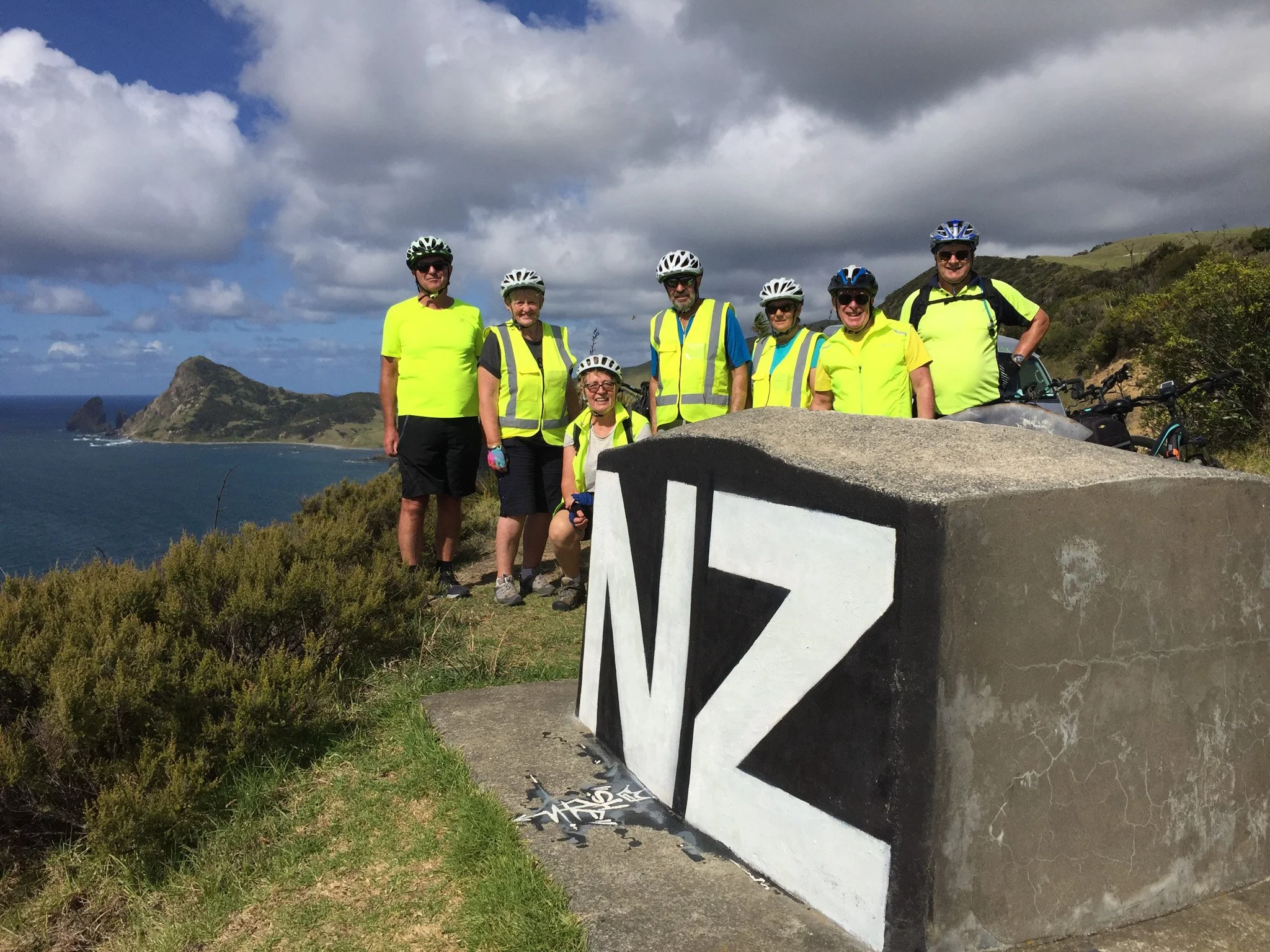 Group of seven people wearing helmets and bright yellow shirts standing on a hilltop overlooking the ocean and mountains under a cloudy sky.