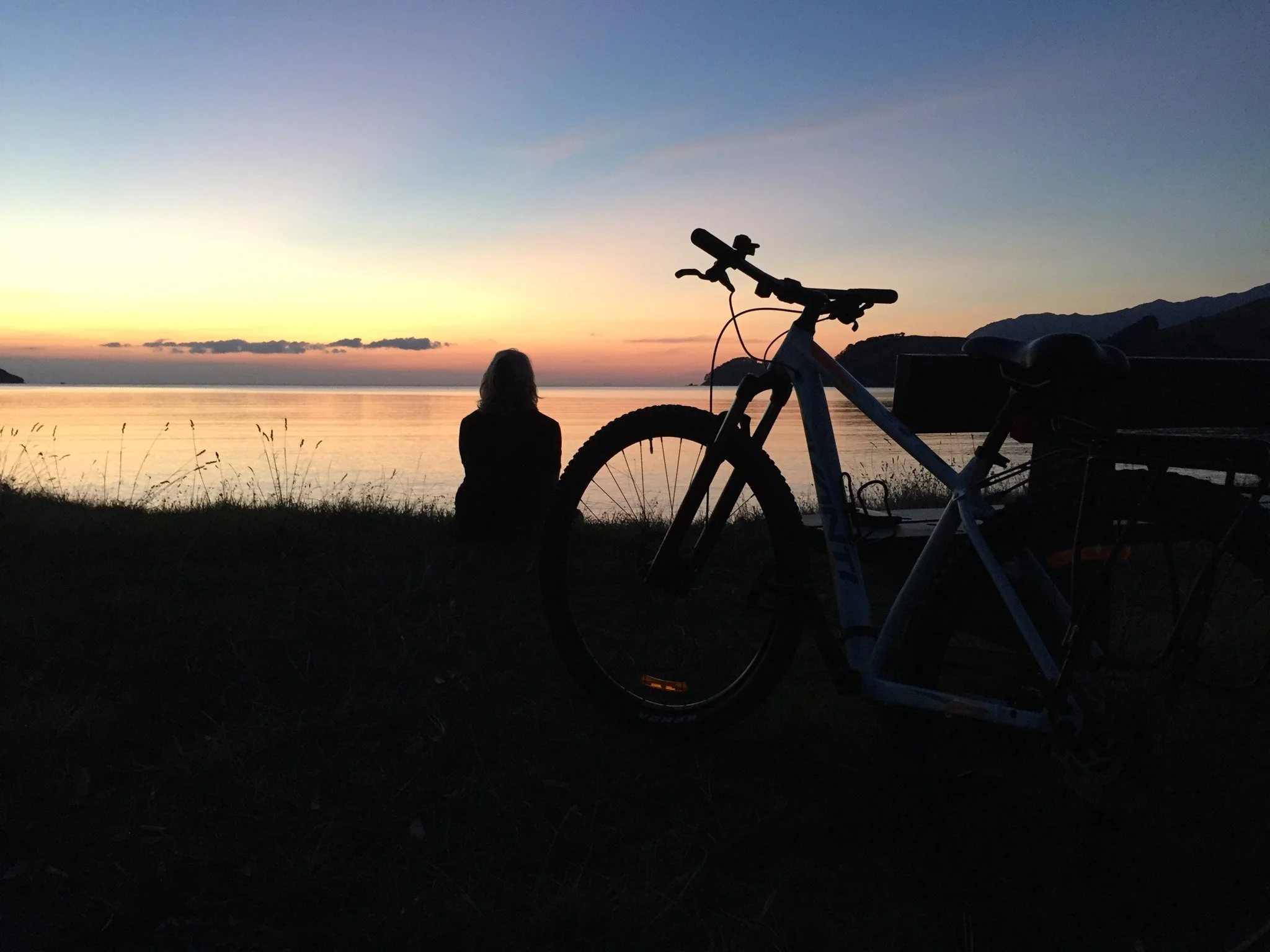 Silhouette of a person sitting beside Colville Bay during sunset, with a bicycle nearby, and distant hills in the background.