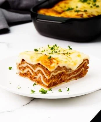 A slice of lasagne on a white plate garnished with fresh herbs, with a baked casserole dish in the background.