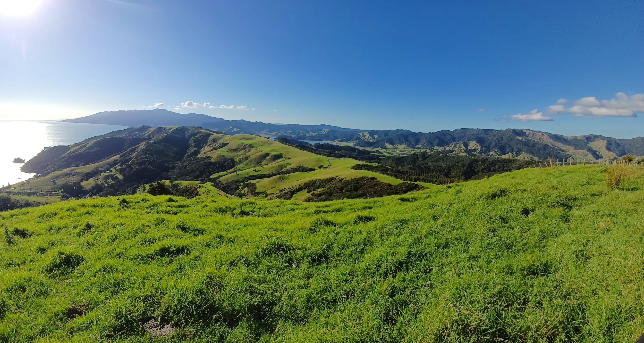 Lush green rolling hills with a coastline to the left, distant mountains under a clear blue sky.