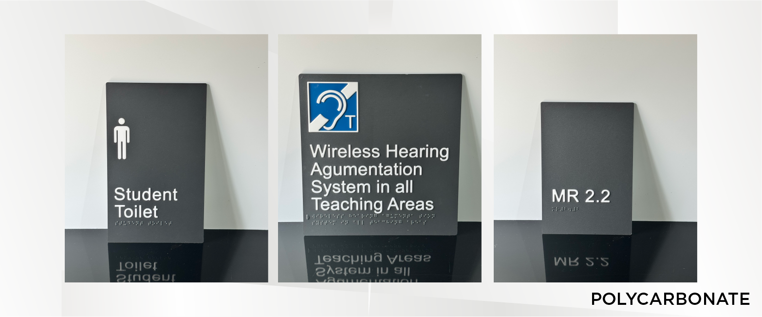 Three black signs with white text displaying directions and information lying on a reflective surface. The first sign points to a student toilet, the second discusses wireless hearing augmentation systems in teaching areas, and the third indicates MR 2.2 room.