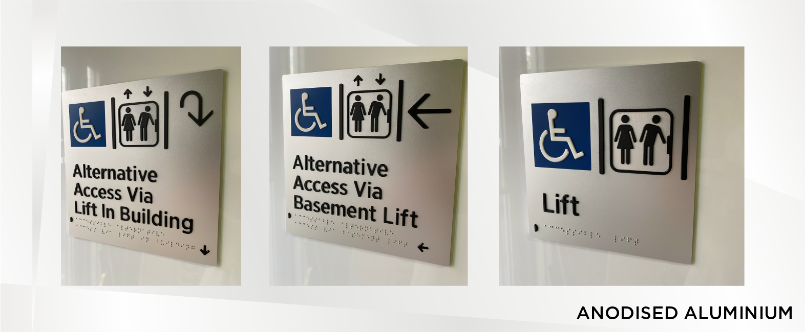 Three signs indicating accessible elevator options. The first sign reads 'Alternative Access Via Lift In Building' with a wheelchair icon and an arrow pointing down. The second sign says 'Alternative Access Via Basement Lift' with a wheelchair icon, male and female figures, and an arrow pointing left. The third sign states 'Lift' with a wheelchair icon and male and female figures. All signs feature braille at the bottom. The signs are made of anodized aluminum.