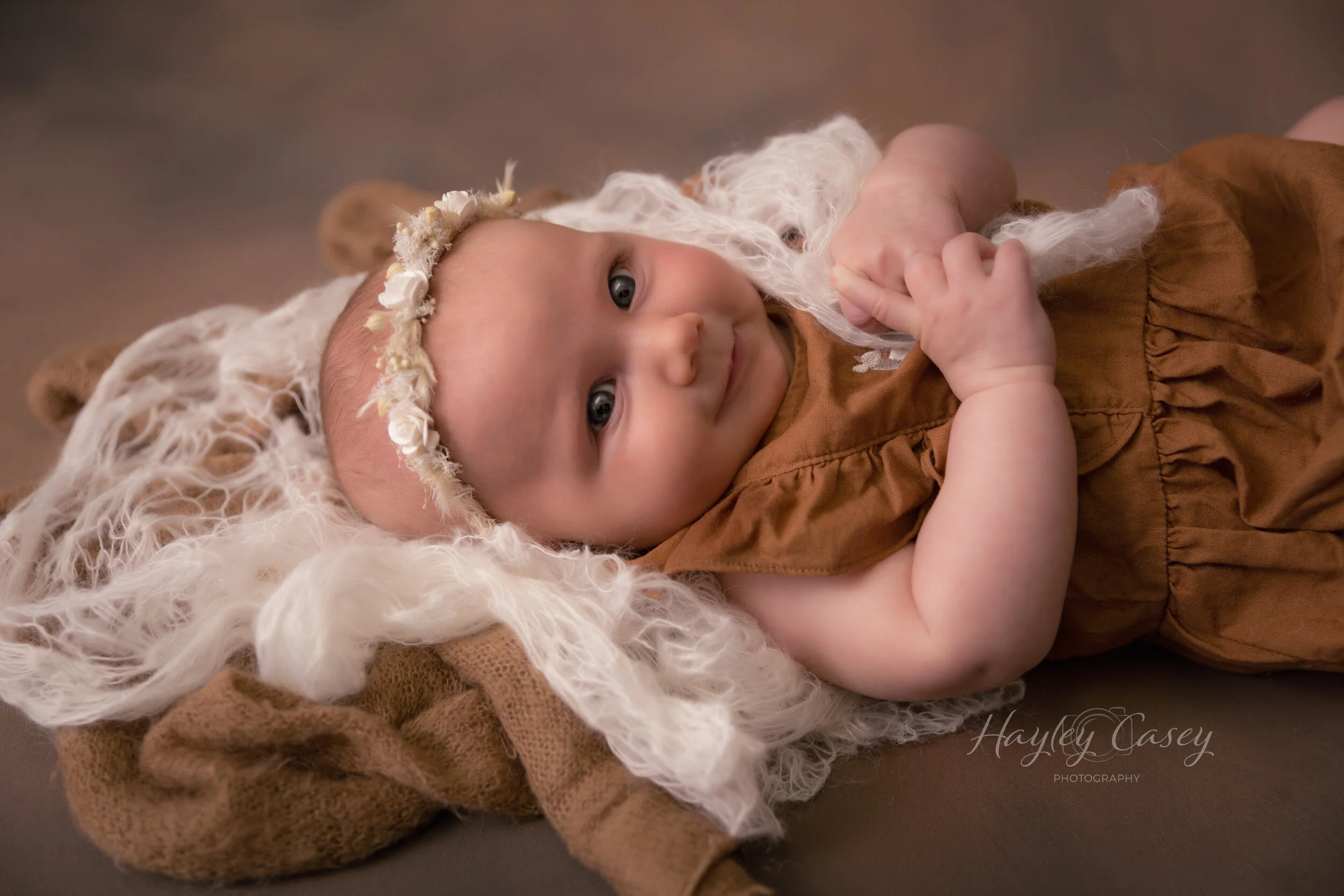 A smiling baby with blue eyes lying on a brown and white animal-shaped blanket, wearing a brown dress and a flower crown.