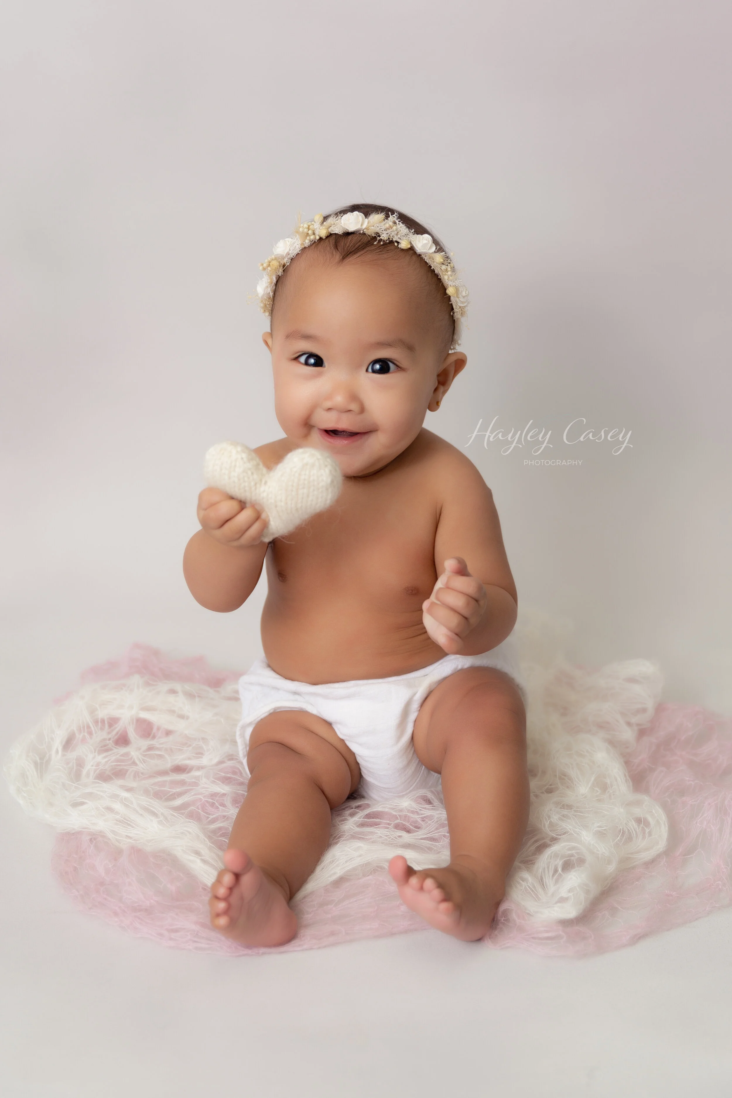 Adorable baby girl sitting on a soft pink and white blanket, wearing a flower headband, white diaper, and holding a small knitted heart, smiling at the camera.