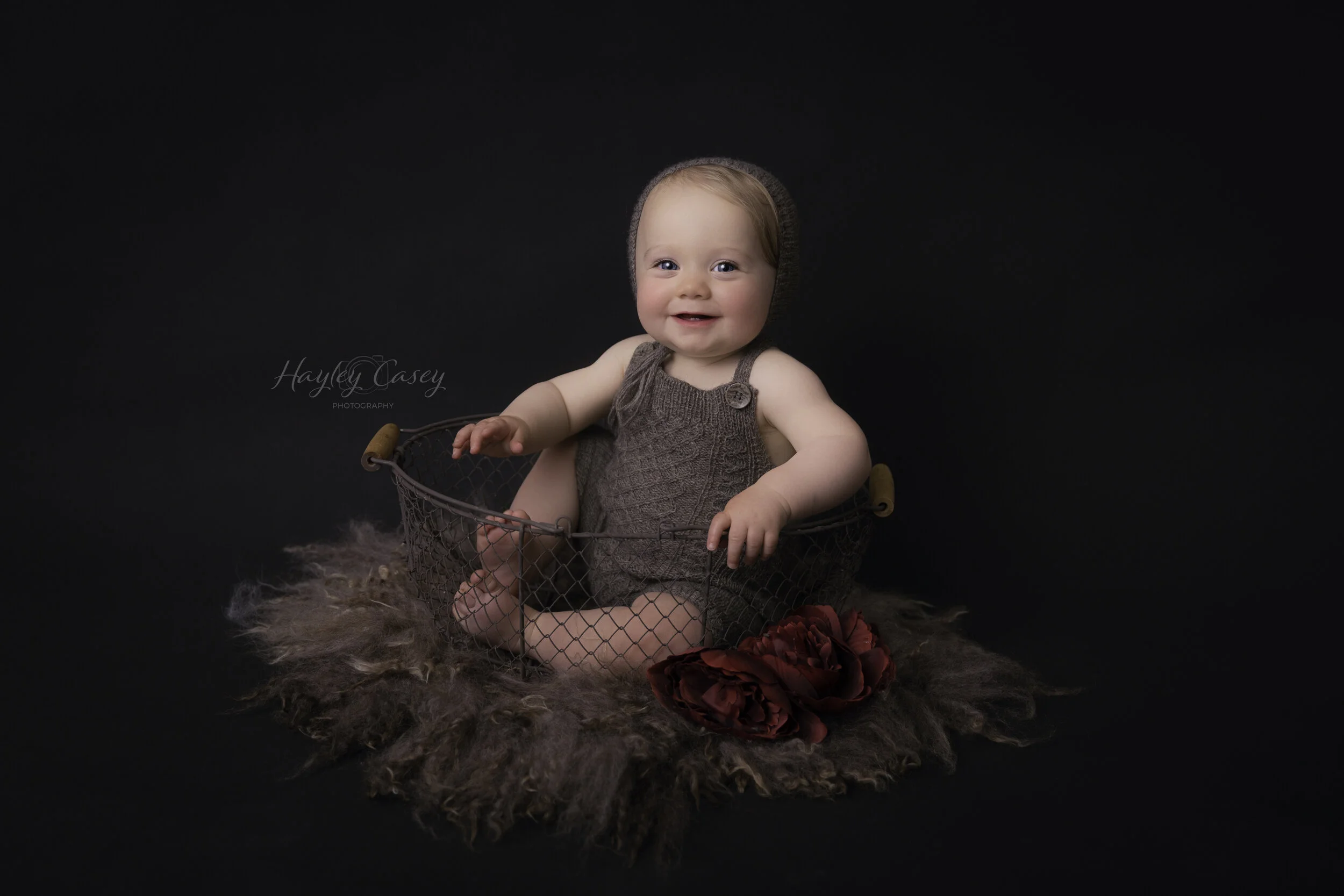 A baby girl sitting in a wire basket, wearing brown knit overalls with a matching hat, surrounded by a furry rug and red flowers against a dark background.