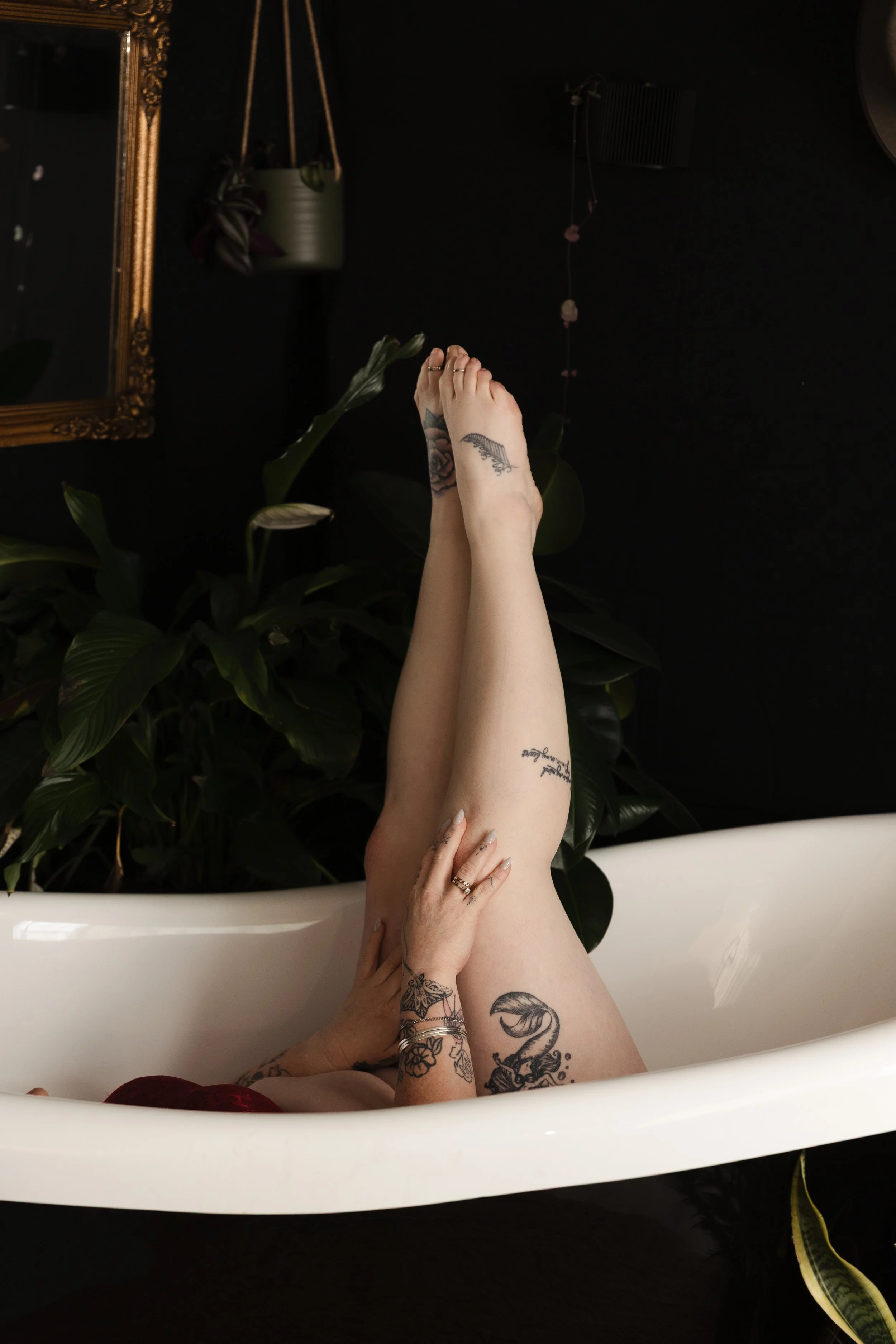 A person with tattoos is relaxing in a white bathtub, with legs raised and crossed, in a dark room with plants, a mirror, and decorative elements.