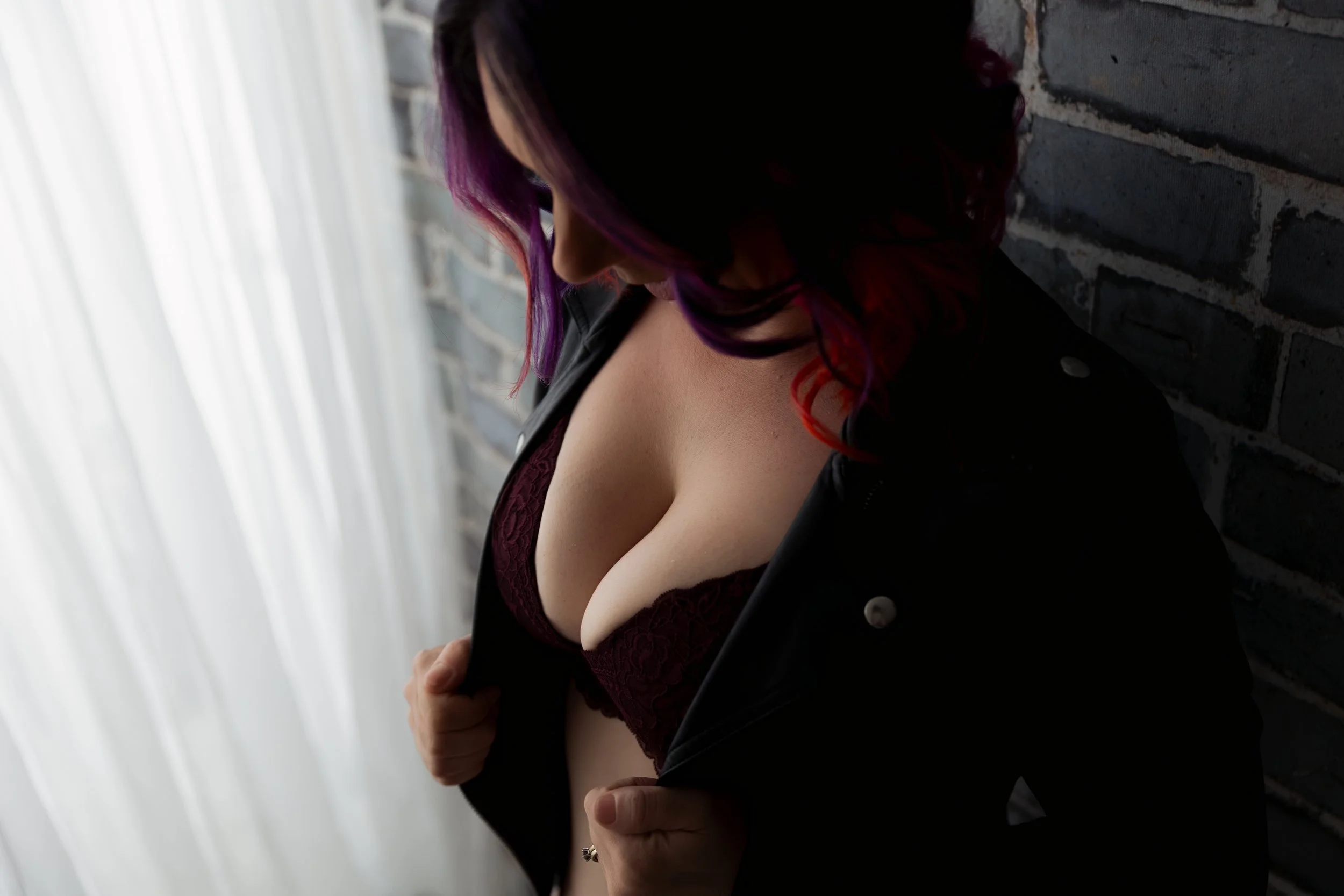 A woman with purple and red hair wearing a maroon lace bra and a black jacket, standing against a brick wall, looking downward. Boudoir photography studio Oamaru, Otago New Zealand 