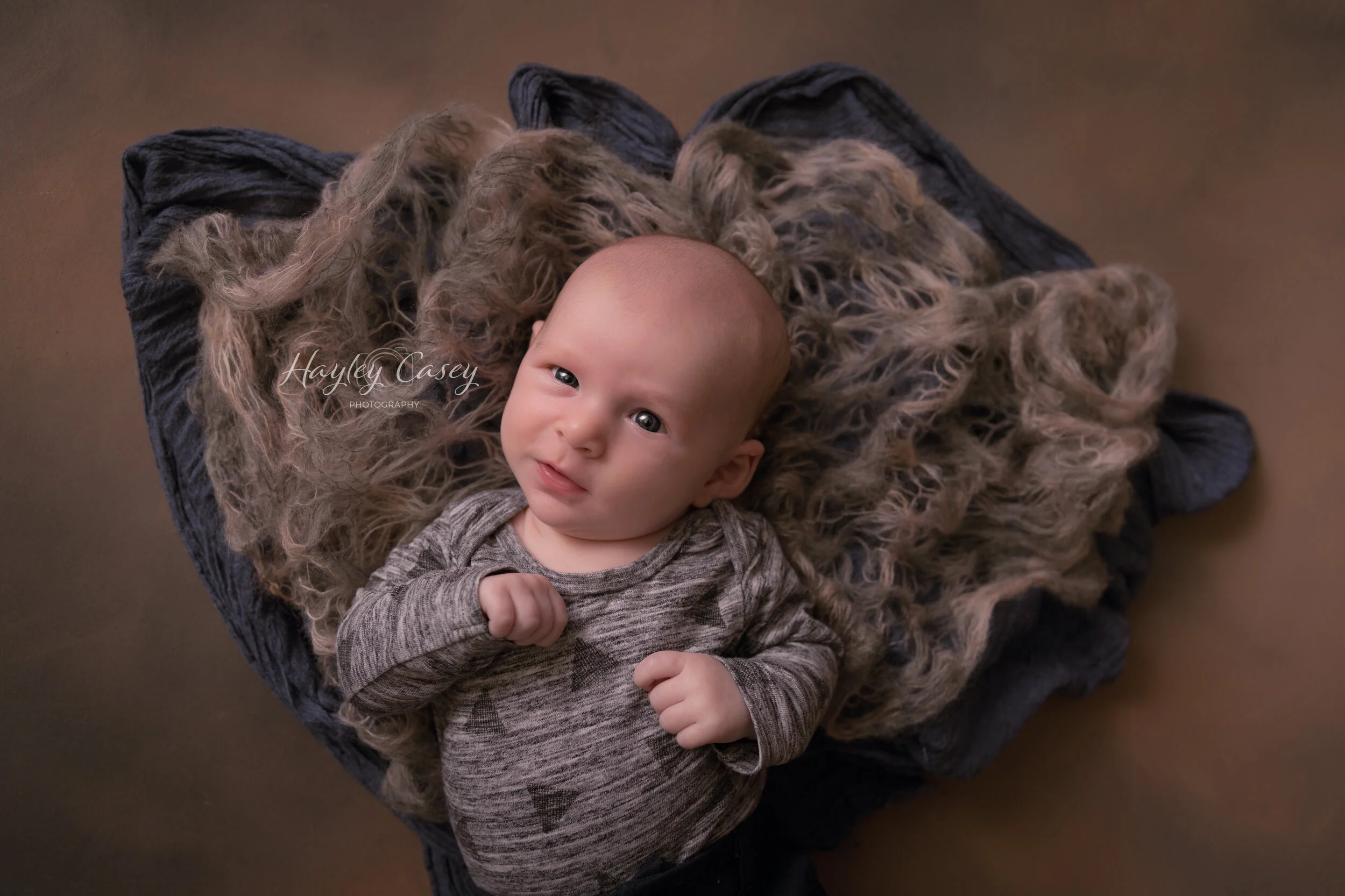 Young baby with light skin, light hair, and bright eyes lying on a dark cushion, surrounded by a soft, curly, beige blanket. The baby is looking up at the camera with a slight smile and wearing a gray, long-sleeved shirt.