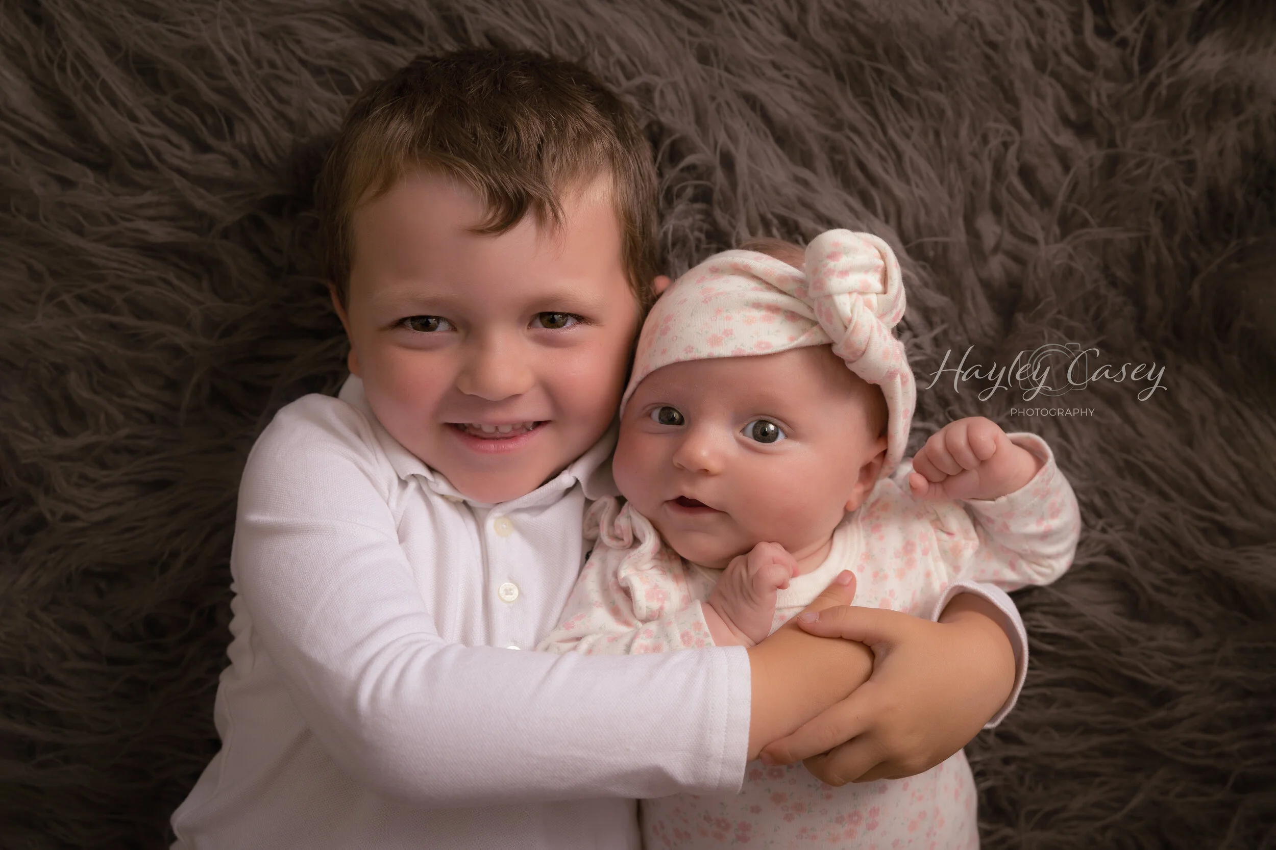 A young boy with brown hair and a white shirt hugs a baby girl wearing a pink and white outfit with a matching headband, lying on a fuzzy brown blanket.
