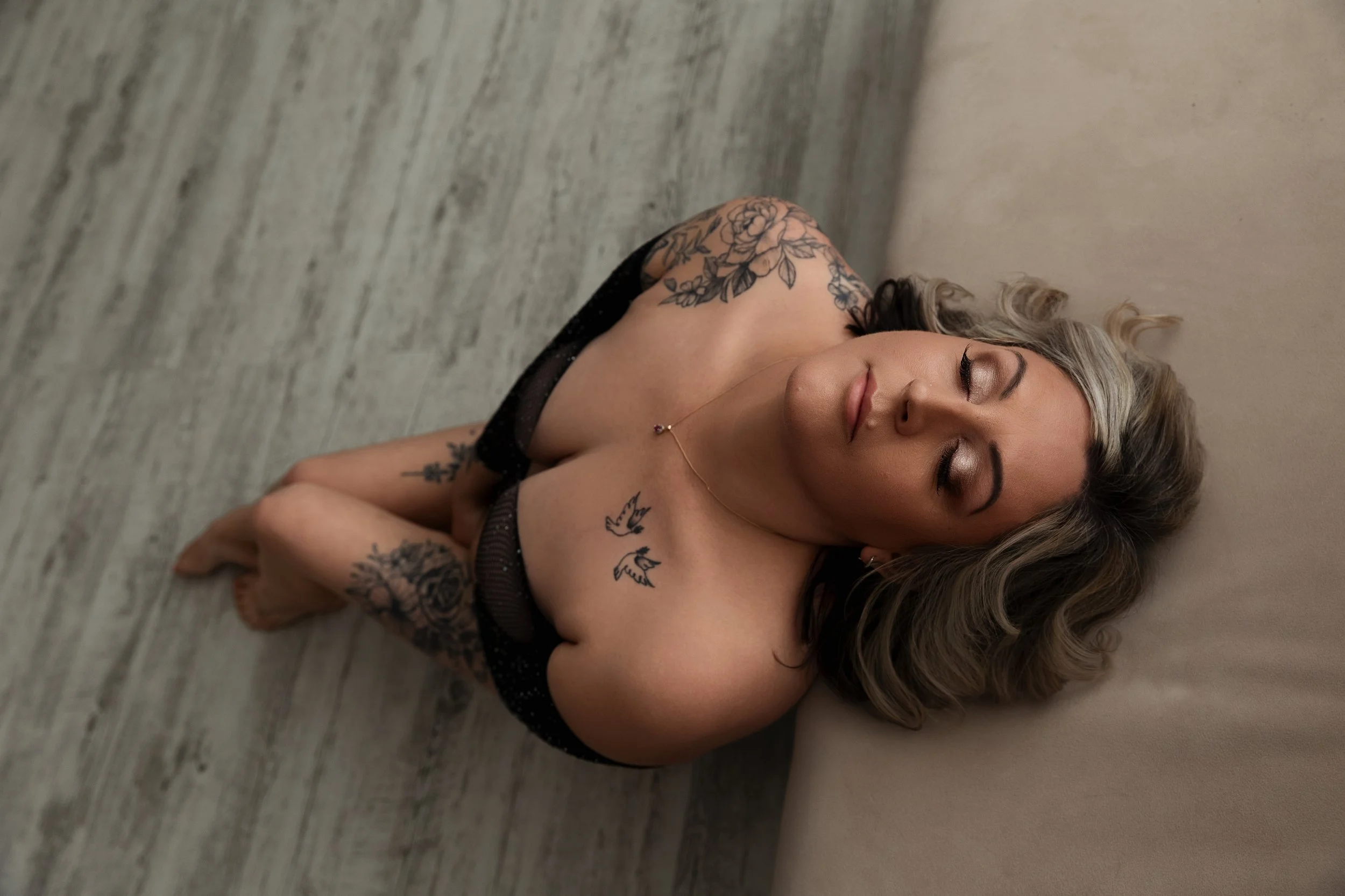 A woman lying on the floor with her eyes closed, wearing a black lace top, with tattoos on her arms and shoulder, and blonde hair with darker roots.