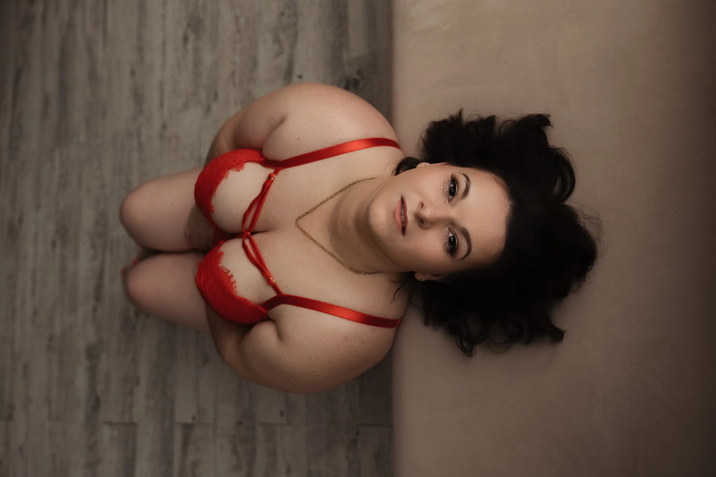 A woman with dark, curly hair lying on the floor, wearing red lingerie and a gold necklace, looking at the camera with a slight smile.