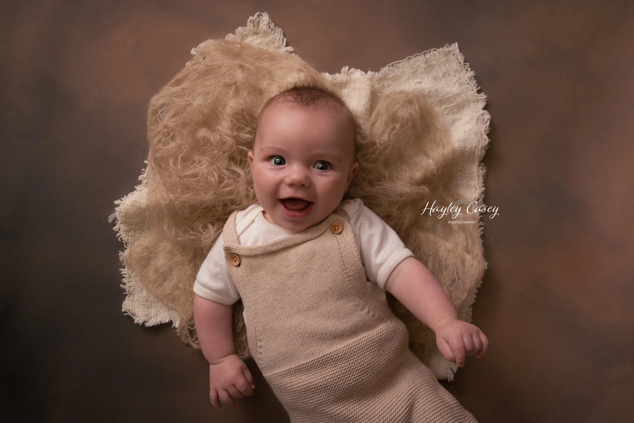 A smiling baby lying on a fluffy beige blanket, wearing a cream-colored knitted jumpsuit with buttons, looking at the camera.