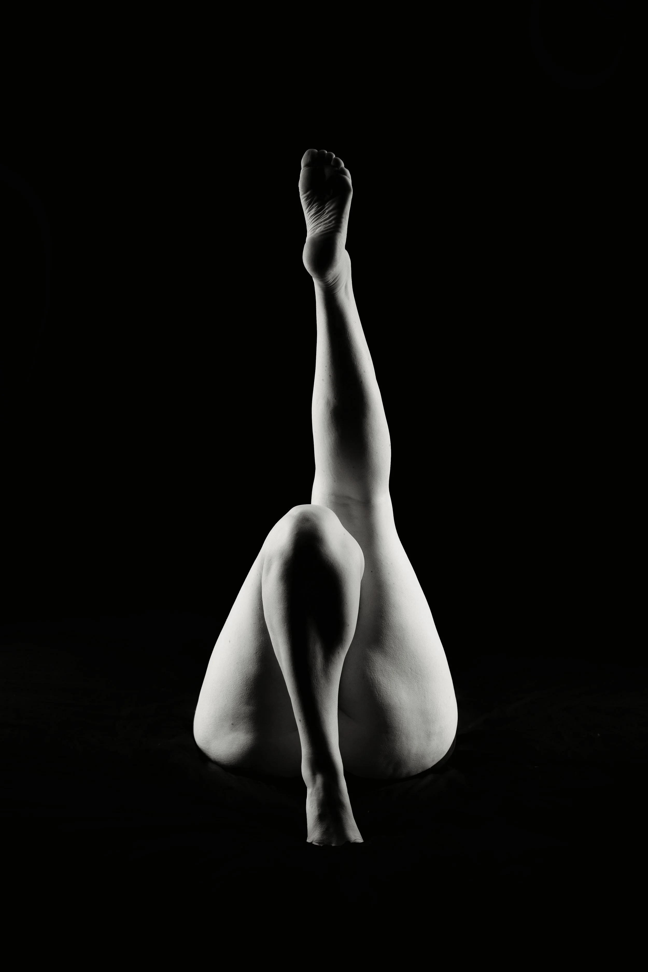 A black and white photograph of a nude person, lying on their back with legs bent and feet raised, against a dark background.